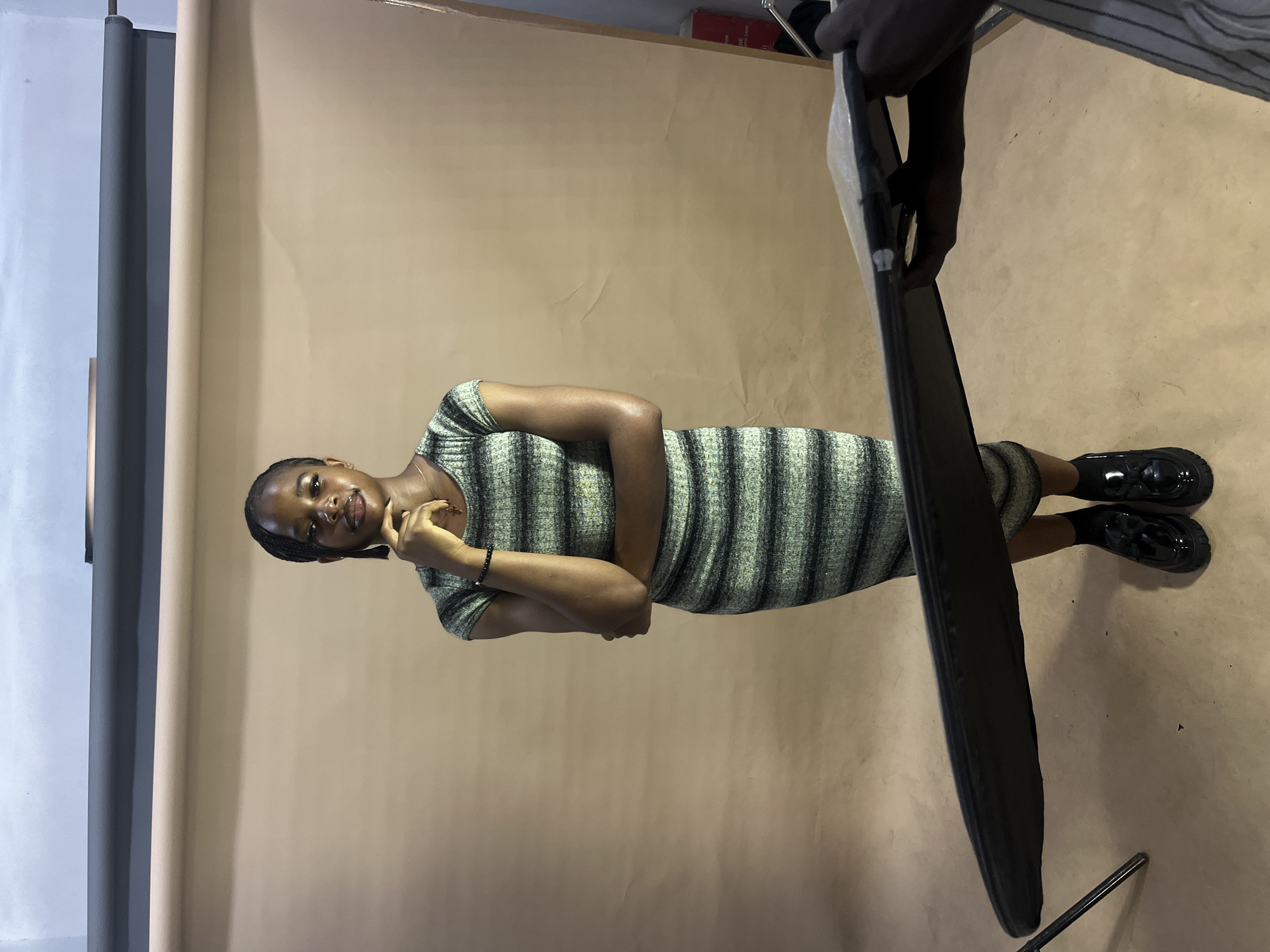 A young woman stands posing in a studio setting. She is wearing a striped, form-fitting dress and black patent leather shoes. Her arms are crossed, and she is looking towards the camera with a slight smile. The background is a plain, light brown paper backdrop. To the right of the woman, a dark, rectangular object, possibly a large photography prop or piece of equipment, is held partially in view by a person whose hand and arm are visible. Another hand, belonging to the same person, is also visible at the top of the frame, holding the object. The lighting appears to be artificial, consistent with a studio environment. No specific time of day or weather conditions are discernible. There is no visible text in the image. The location context is Gura Top, Nigeria.