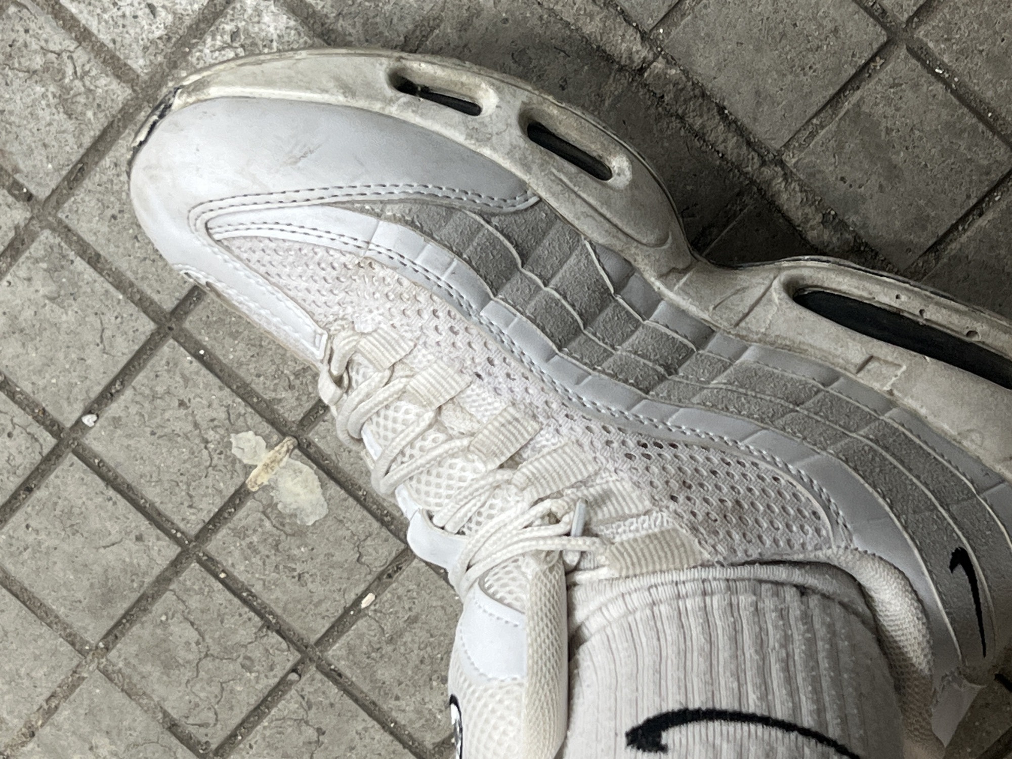 A worn, white and grey Nike Air Max 95 athletic shoe is positioned on a cracked, grey, square-tiled concrete surface in Debelt, Bulgaria. The left foot, encased in the shoe, wears a white ribbed sock with a prominent black Nike swoosh logo on its outer side. The shoe's white mesh upper and grey layered side panels exhibit visible dirt and scuff marks. The white midsole, which contains dark, recessed air units, also shows signs of wear and dirt accumulation. The toe cap area appears discolored or scuffed. The ground features an irregular pattern of cracks and debris, including several small white fragments and a larger, irregularly shaped piece of light-colored material located near the front of the shoe.