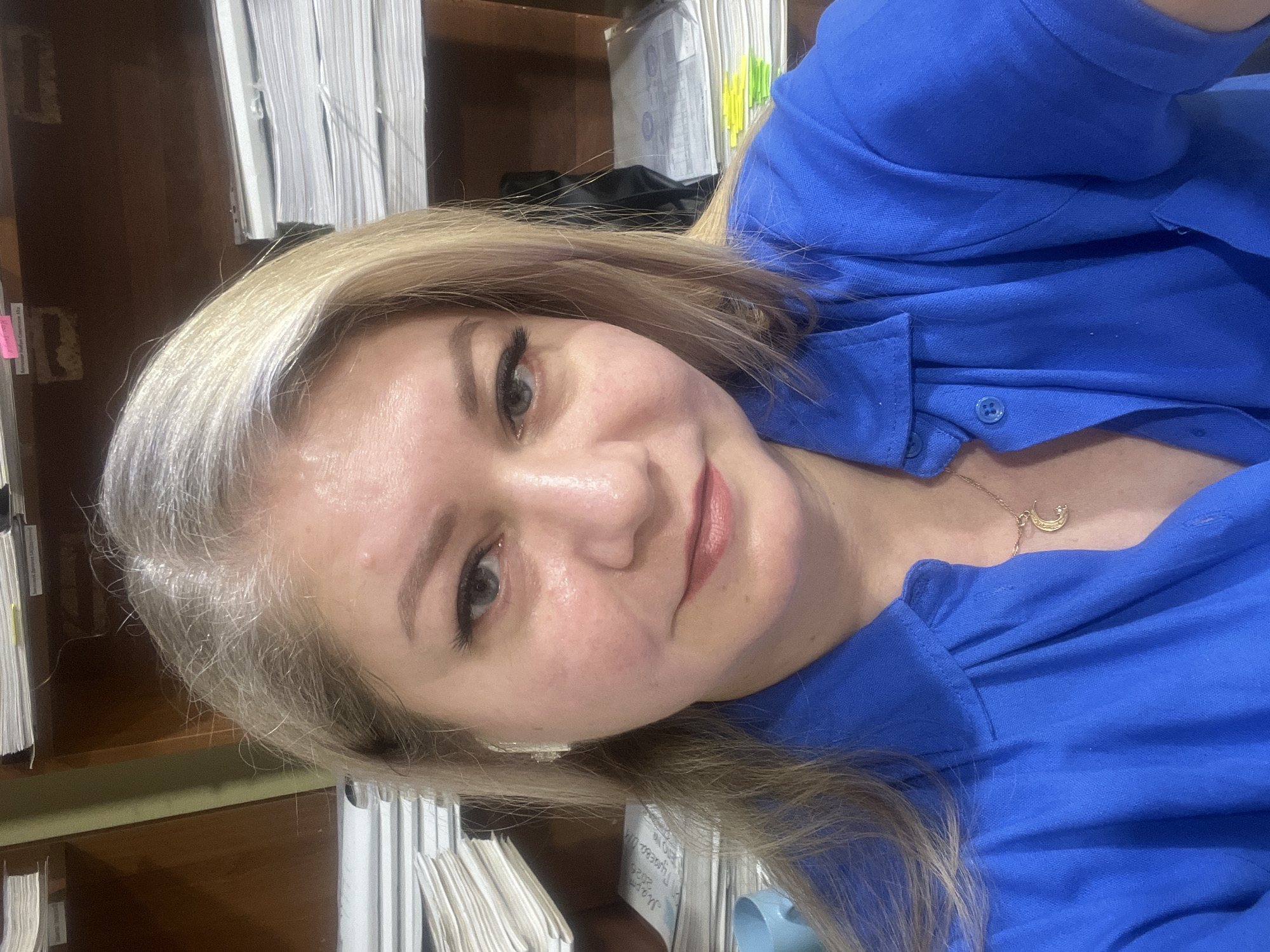 A woman with blonde hair is posing for a selfie in what appears to be an office. She is wearing a bright blue collared shirt and a delicate gold necklace with a crescent moon pendant. Behind her, shelves are stacked with piles of white papers. The location in Izhevsk, Russia cannot be determined from the image.