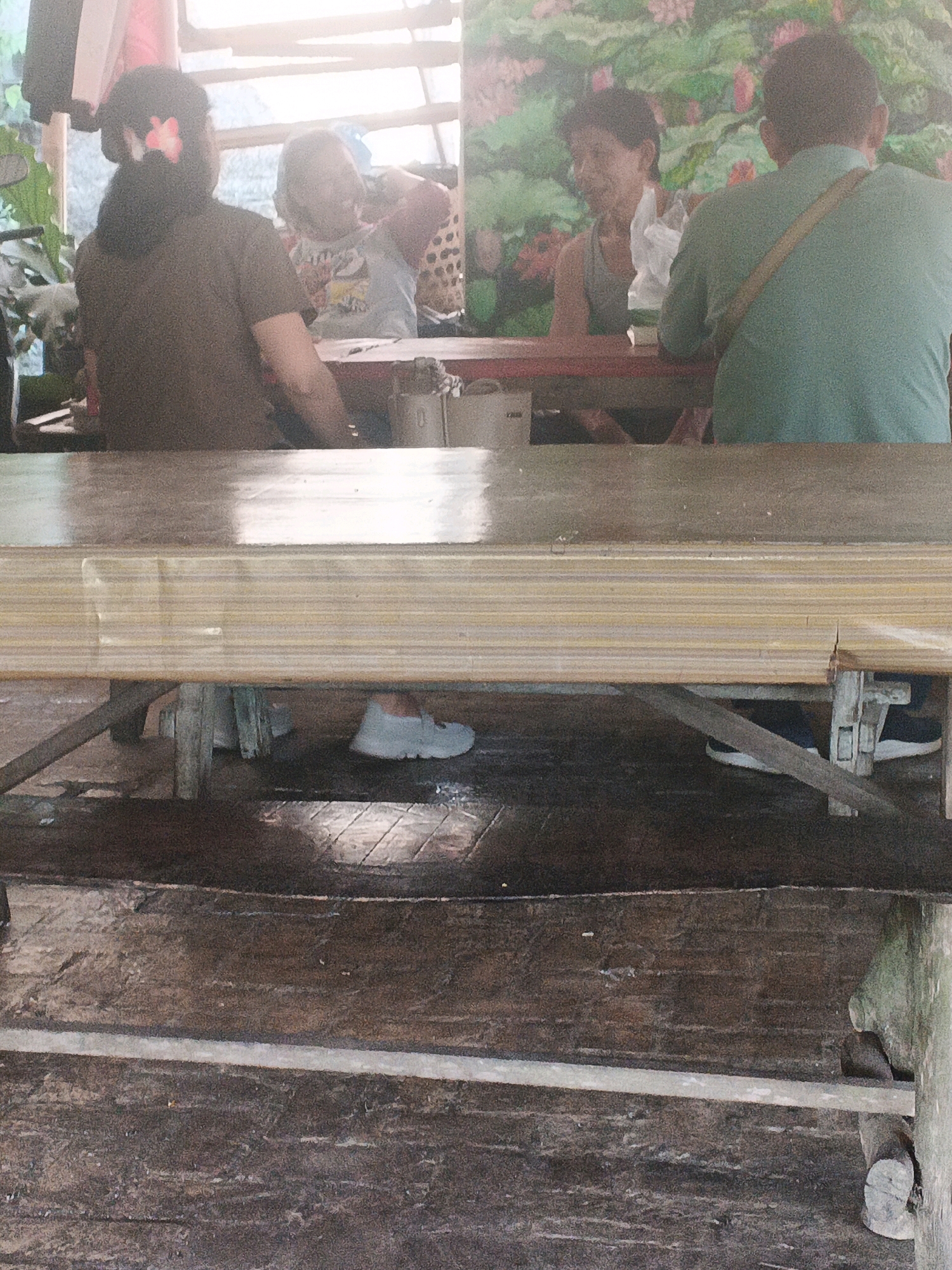 In Ungaran, Indonesia, four individuals are seated at a table in what appears to be an outdoor or semi-outdoor setting, possibly a warung or food stall. The overall atmosphere is casual.
In the foreground, a light-colored wooden table is positioned horizontally across the frame, with a darker wooden floor beneath it. The floorboards are worn and show some signs of dirt and moisture.
The people are visible from the mid-section upwards. On the left, a woman with dark hair, adorned with a pink flower, is facing away from the camera, wearing a dark t-shirt. To her right, a child is seated, looking towards the left. Further to the right, a man in a grey tank top is looking towards the center, holding a white plastic bag. On the far right, a man in a green t-shirt is facing away from the camera, with a light-colored strap across his shoulder.
Behind the individuals, a backdrop features a painting of large, green leaves and pink flowers, suggesting a decorative element. The lighting is bright, with sunlight possibly filtering in from above. Some elements of the structure, like wooden beams and possibly corrugated roofing, are visible. On the table, there are some objects that are indistinct but could be food containers or utensils. A white shoe is visible beneath the table on the left side.
