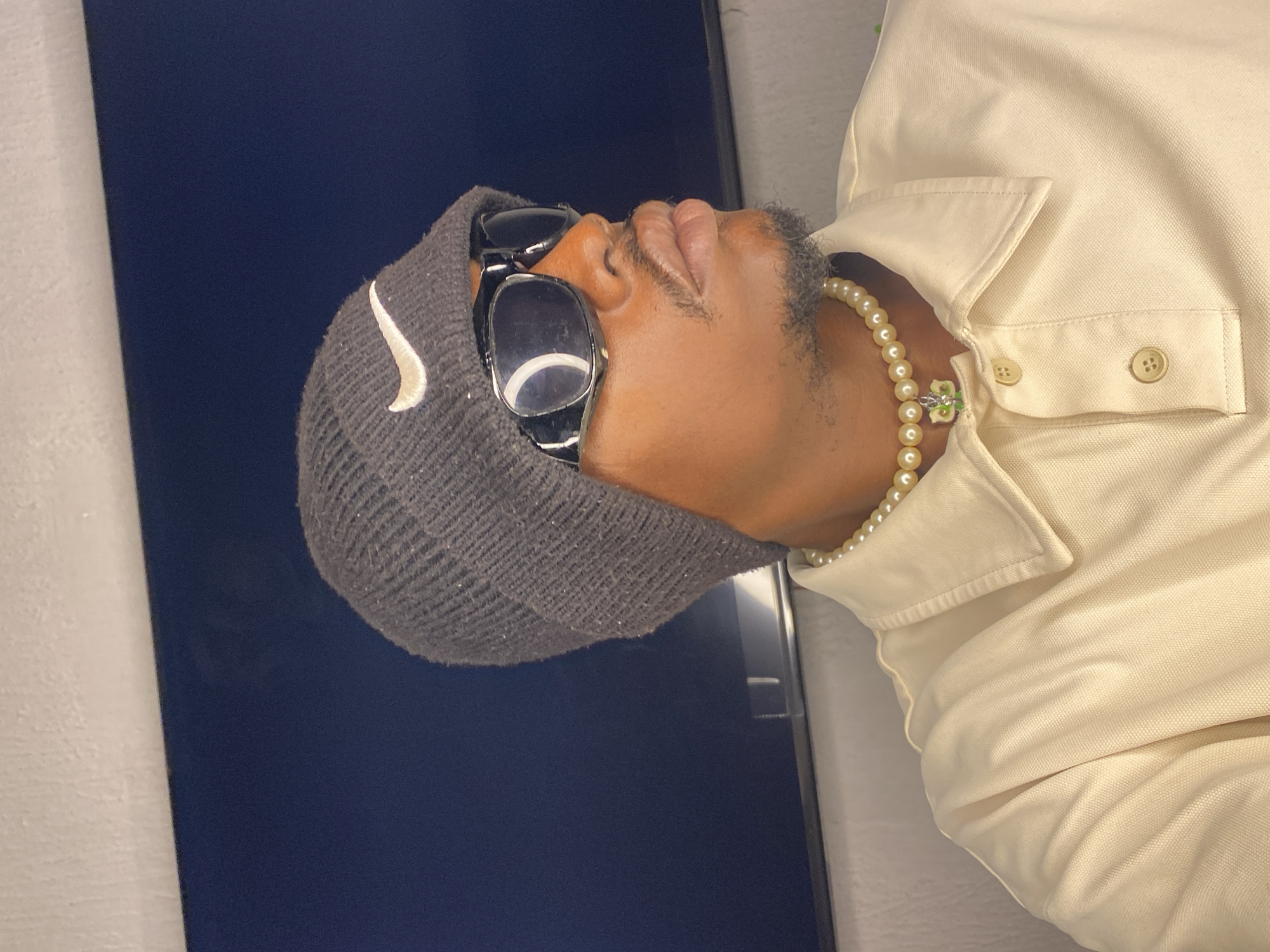 A medium shot shows a person facing to the right, wearing a dark grey beanie with a white Nike logo, black wraparound sunglasses, a pearl necklace with a green and white charm, and a light cream collared shirt. The person has a mustache and goatee and their skin tone is medium brown. They appear to be indoors, in front of a dark blue surface, possibly a screen or wall, with a lighter, textured surface above. The lighting is soft, creating shadows on the person's face and the right side of their body. The scene is devoid of any recognizable urban or natural landmarks that would pinpoint a specific location within Abakaliki, Nigeria.