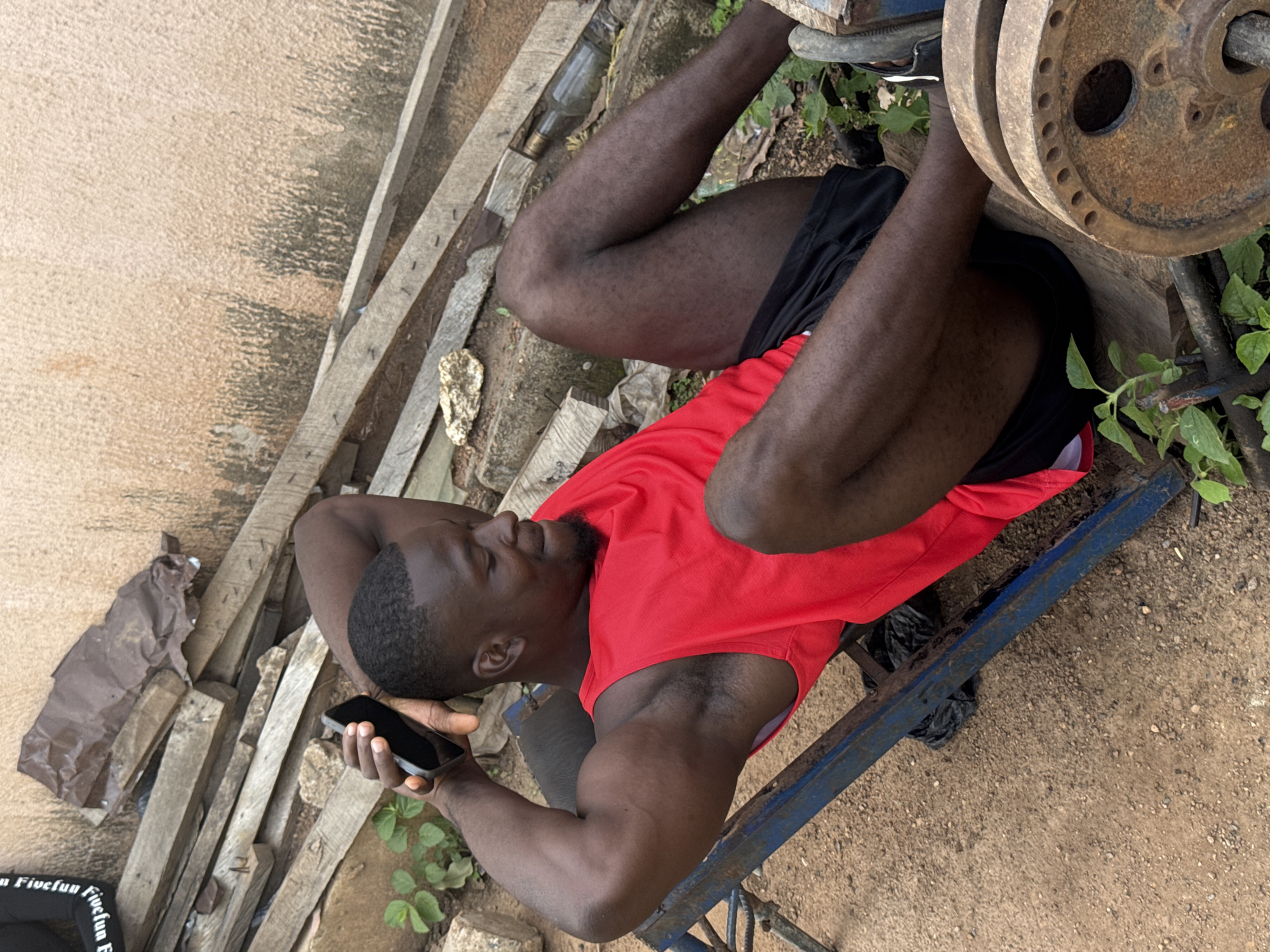 A man, appearing to be of African descent, is lying down on a workout bench with a barbell resting on his feet. He is wearing a red tank top and black shorts. The man is holding a smartphone in his left hand and looking down at it. The scene appears to be outdoors, with a dirt ground and some scattered wooden planks. There is also some greenery visible, suggesting a natural setting. The lighting suggests it is daytime. A rusty weight plate is visible on the barbell. Some debris, including what looks like a plastic bag and a glass bottle, are scattered around the scene. The man's facial expression is neutral, possibly focused on his phone. Some text is visible on a black fabric item in the lower left corner of the image.