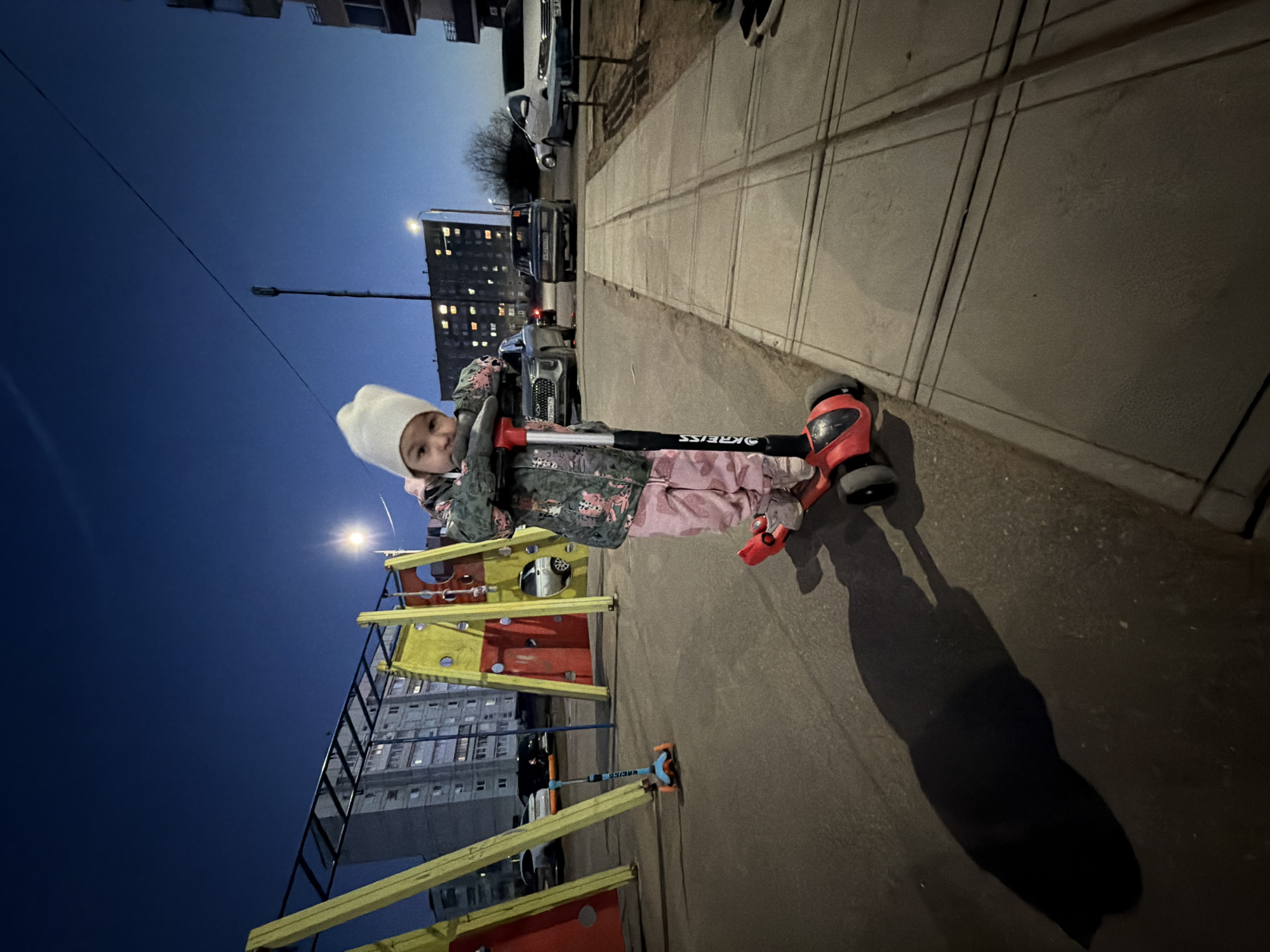 A young child in a white hat, pink pants, and a camouflage jacket is riding a scooter on a sidewalk near a playground in Chaikovsky, Russia. The sky is dusky, suggesting early evening. Behind the child and playground are tall apartment buildings with lit windows. Cars are parked along the road, and streetlights illuminate the scene. The sidewalk is made of large, square paving stones. The scooter is red and black with the brand "STELS" visible. Another, smaller scooter is visible near the playground equipment. The image appears to be taken from a low angle.