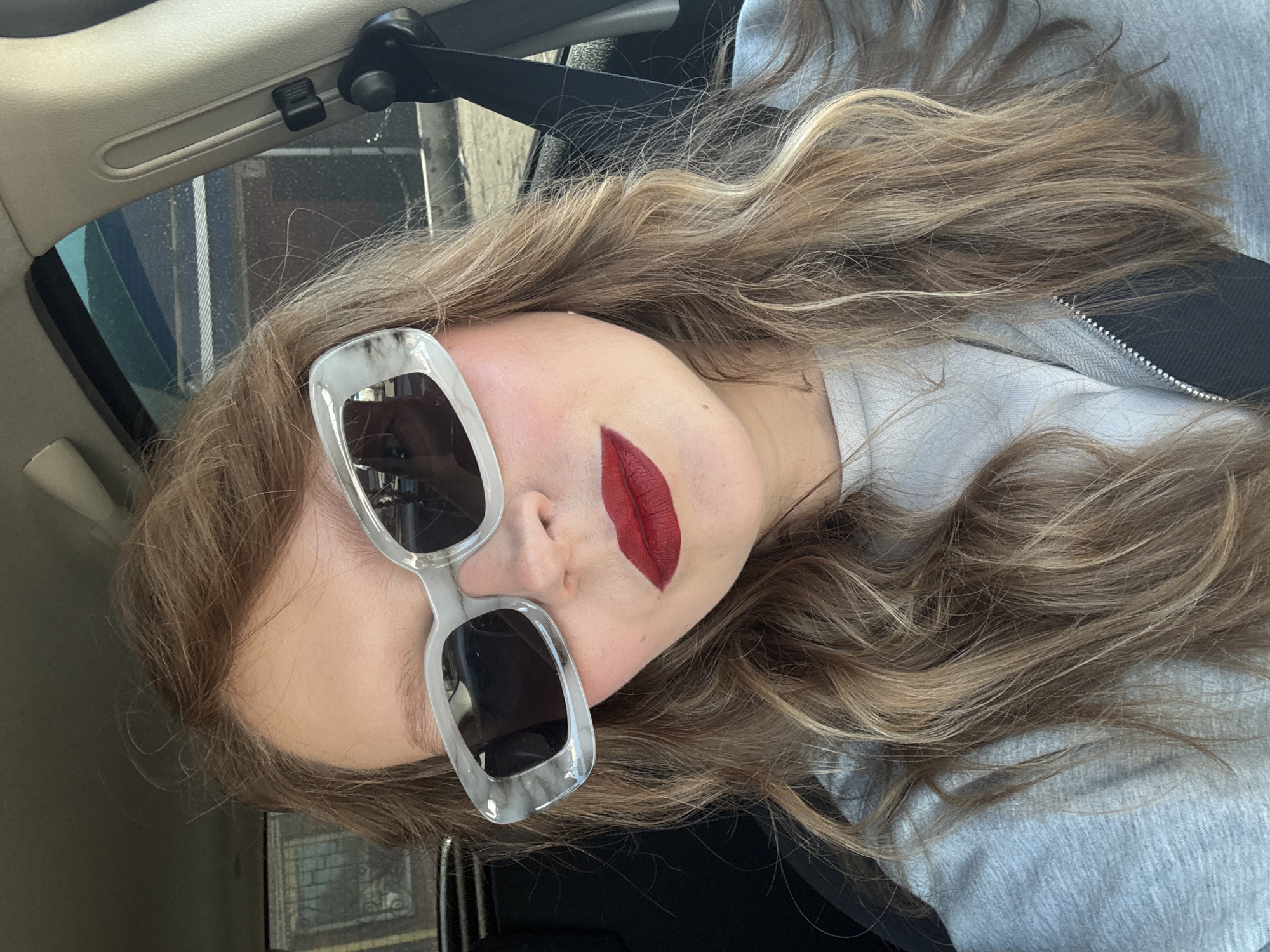 A medium shot selfie shows a young woman in a car, wearing large, light-colored sunglasses with a marble-like pattern. Her hair is long and wavy, a mix of blonde and brown tones. She is wearing a grey zip-up hoodie over a lighter grey top. Her lips are painted a bold red. The background shows the interior of a car, with a seatbelt visible. Through the car window, parts of buildings and trees can be seen, suggesting an urban environment, possibly Aksay, Kazakhstan.