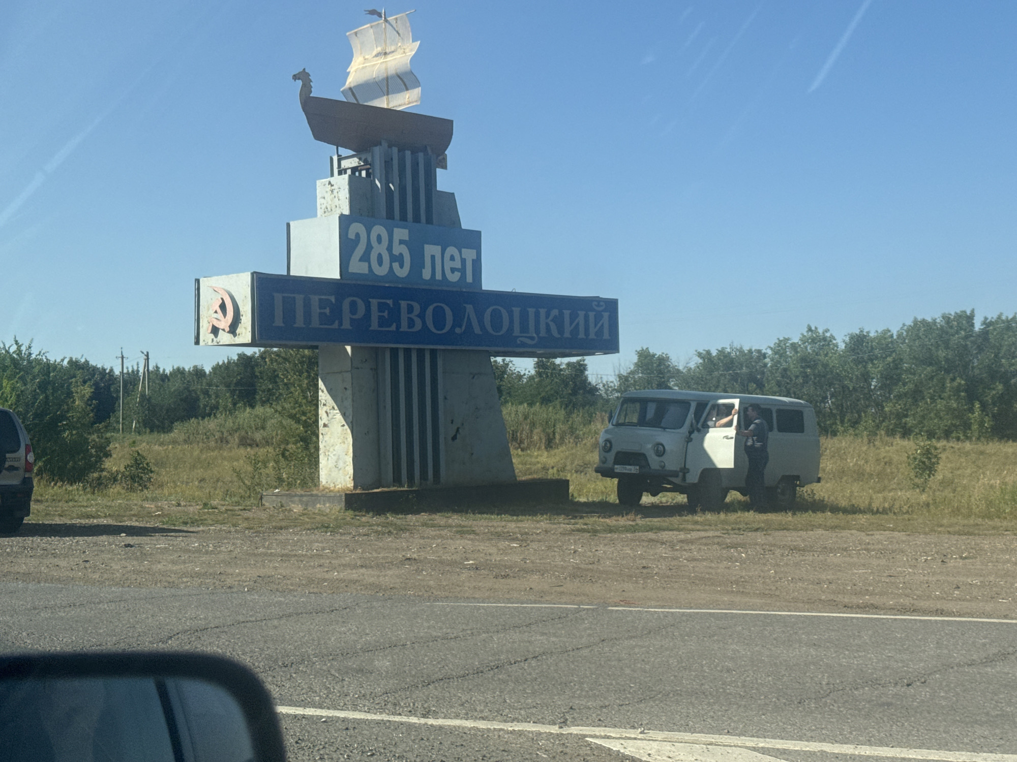 A monument in Perevolotsky, Russia, commemorates the town's 285th anniversary. The monument features a Soviet hammer and sickle emblem and a ship with sails. A white van is parked near the monument, and a person is visible inside the van. The scene is set against a backdrop of trees and a clear blue sky.