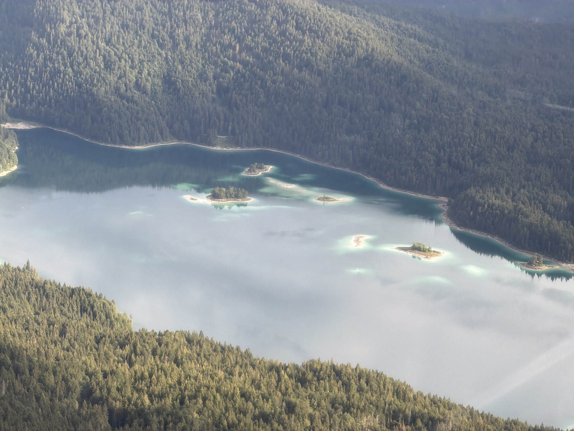 This is an aerial, high-angle photograph of a body of water, likely a lake, surrounded by dense coniferous forests. The water is a bright turquoise or teal color in shallower areas, transitioning to a deeper blue where it is more profound. Several small, tree-covered islands are scattered across the surface of the water. The shoreline is defined by the dark green of the dense forest that climbs up steep hillsides. The overall impression is one of a natural, serene landscape. The image is a view of the Eibsee lake, located in Grainau, Germany, at the foot of the Zugspitze mountain. There are no people or man-made structures visible in the image, only the natural elements of water and forest.