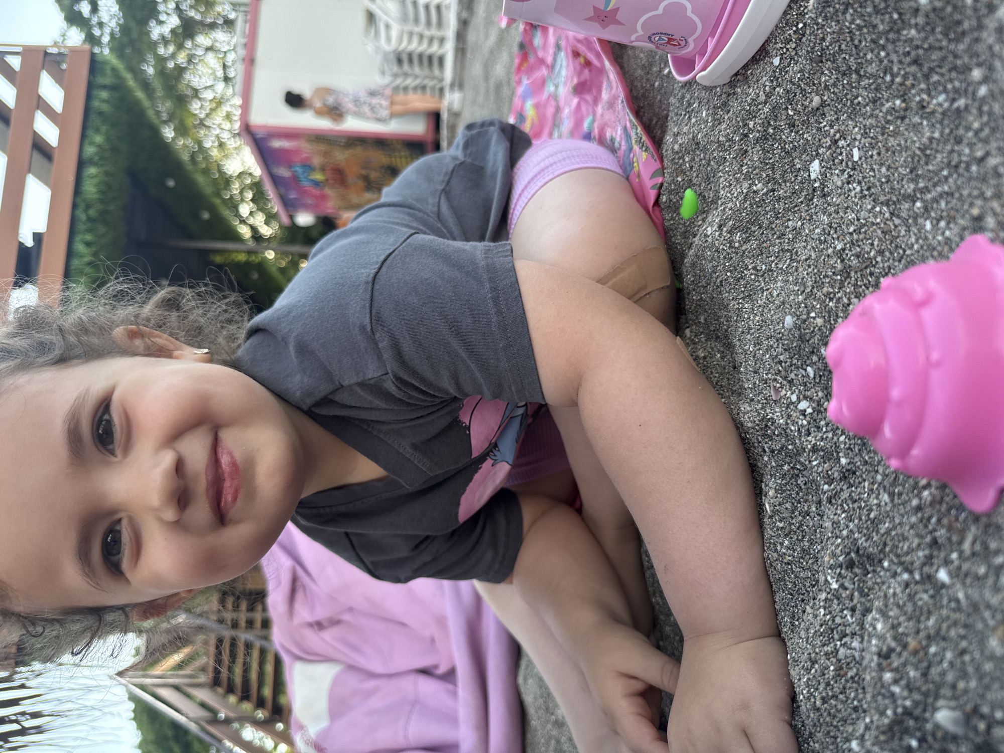 The image shows a close-up of a young girl with curly hair sitting on dark sand, likely at a beach in Burgas, Bulgaria. She is wearing a dark gray t-shirt with a pink graphic and purple shorts. Her legs are crossed, and she is looking towards the camera with a smile. To her right, there is a pink sand mold and a pink bucket. In the background, there is a pink blanket, wooden stairs, a green hedge, and a white building with colorful murals, suggesting a beach resort or a similar recreational area. Two other figures, one adult and one child, are visible in the distance. The lighting indicates it is daytime, possibly late afternoon due to the warm tones.