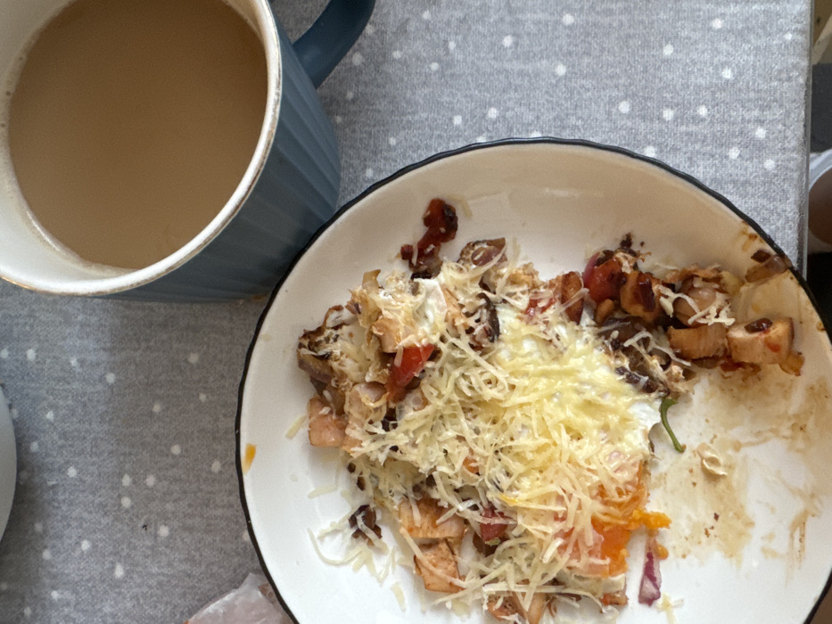 This image depicts a partially consumed meal and a beverage situated on a patterned surface in an indoor setting in Odintsovo, Russia. The primary subject is a white ceramic plate with a dark rim, containing food that includes a fried egg with a visible orange yolk, shredded light-colored cheese, diced meat pieces (possibly chicken or bacon), and fragments of red vegetables (likely tomato or red onion) and darker, caramelized elements. The food shows signs of being partially eaten. To the upper right, a dark teal or blue-green ribbed ceramic mug, accented with a lighter rim, holds a light brown liquid, consistent with coffee or tea with milk. Both the plate and the mug rest on a light grey fabric surface adorned with small, evenly spaced white polka dots. No individuals are visible.