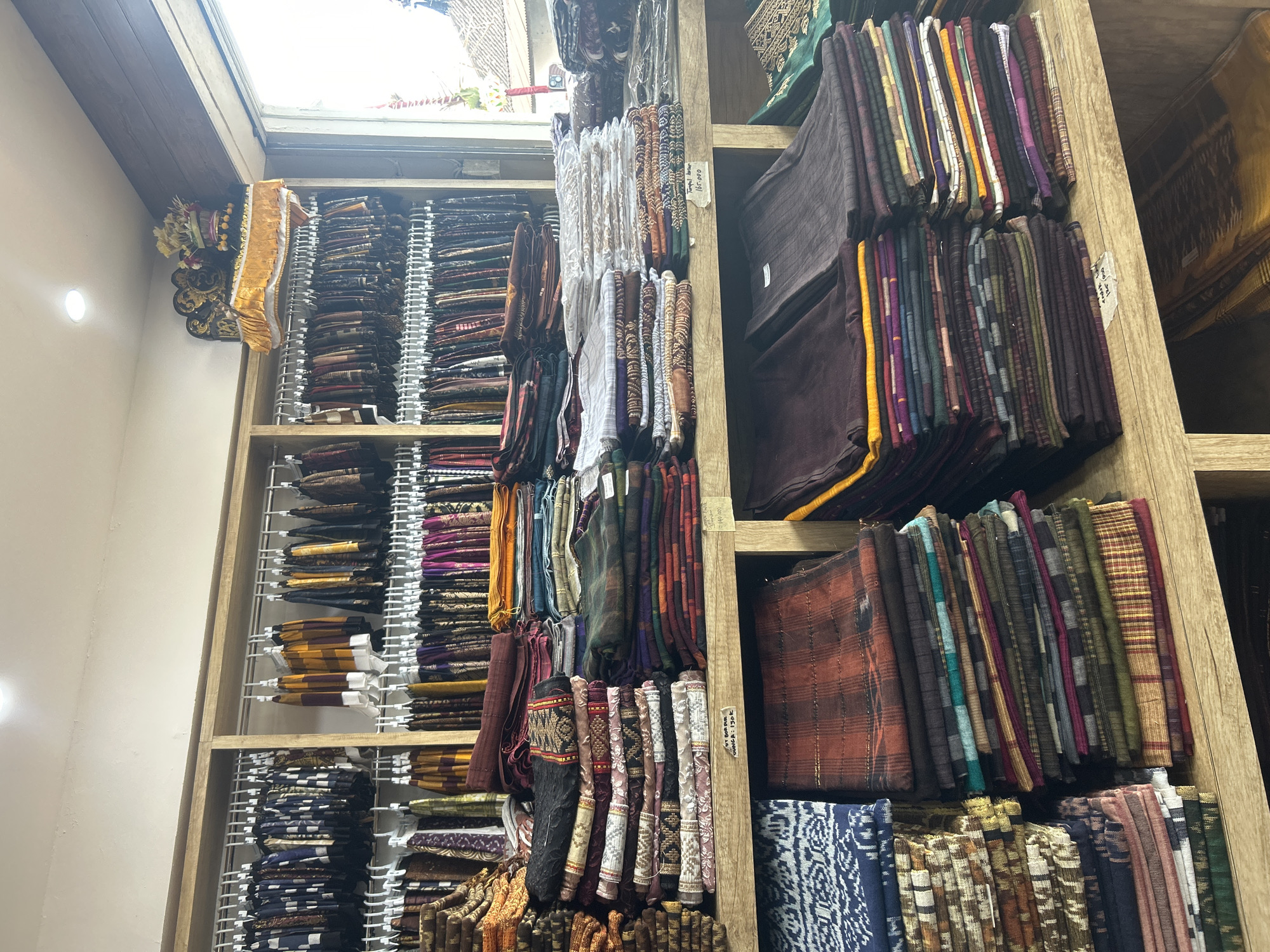 This media file shows a well-organized display of textiles in a shop, likely in Denpasar, Indonesia. The primary subjects are the various fabrics, neatly folded and stacked on wooden shelves. The shelves are filled with a wide assortment of colorful and intricately patterned textiles, including what appear to be sarongs or similar garments. The fabrics display a range of colors, from deep purples and browns to vibrant oranges, yellows, and greens, with many featuring complex geometric and traditional motifs. Some fabrics are hanging, showcasing their texture and flow.
One prominent object is a decorative golden sash or banner hanging from the top left, adorned with what looks like floral arrangements, suggesting a festive or ceremonial context. There are also labels or tags visible on some of the stacks of fabric, indicating potential pricing or product information.
The lighting suggests an indoor setting, possibly during the daytime, with natural light filtering in from above, creating a bright and airy atmosphere. The wooden shelves and the overall presentation suggest a traditional marketplace or boutique specializing in Indonesian textiles. No people are visible in the frame.
Visible text includes a handwritten price tag on one of the fabrics, reading "140.000" and another partially visible tag that appears to say "Batik Tulis / 160.000". This text indicates that the items are likely batik fabrics and provides their prices. The overall impression is one of abundance and craftsmanship, highlighting the rich textile heritage of the region.