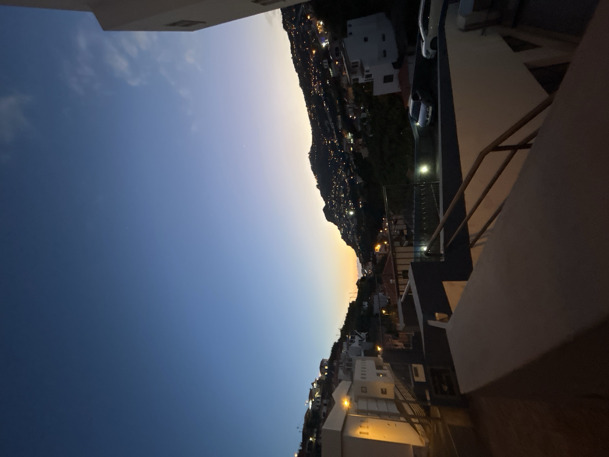 The image shows a view of Câmara de Lobos, Portugal, at twilight. The sky transitions from blue to a lighter shade near the horizon, where it meets a hill covered in buildings with illuminated windows. The perspective is from an elevated position, possibly a balcony, with railing and building elements in the foreground. A couple of cars are also visible.