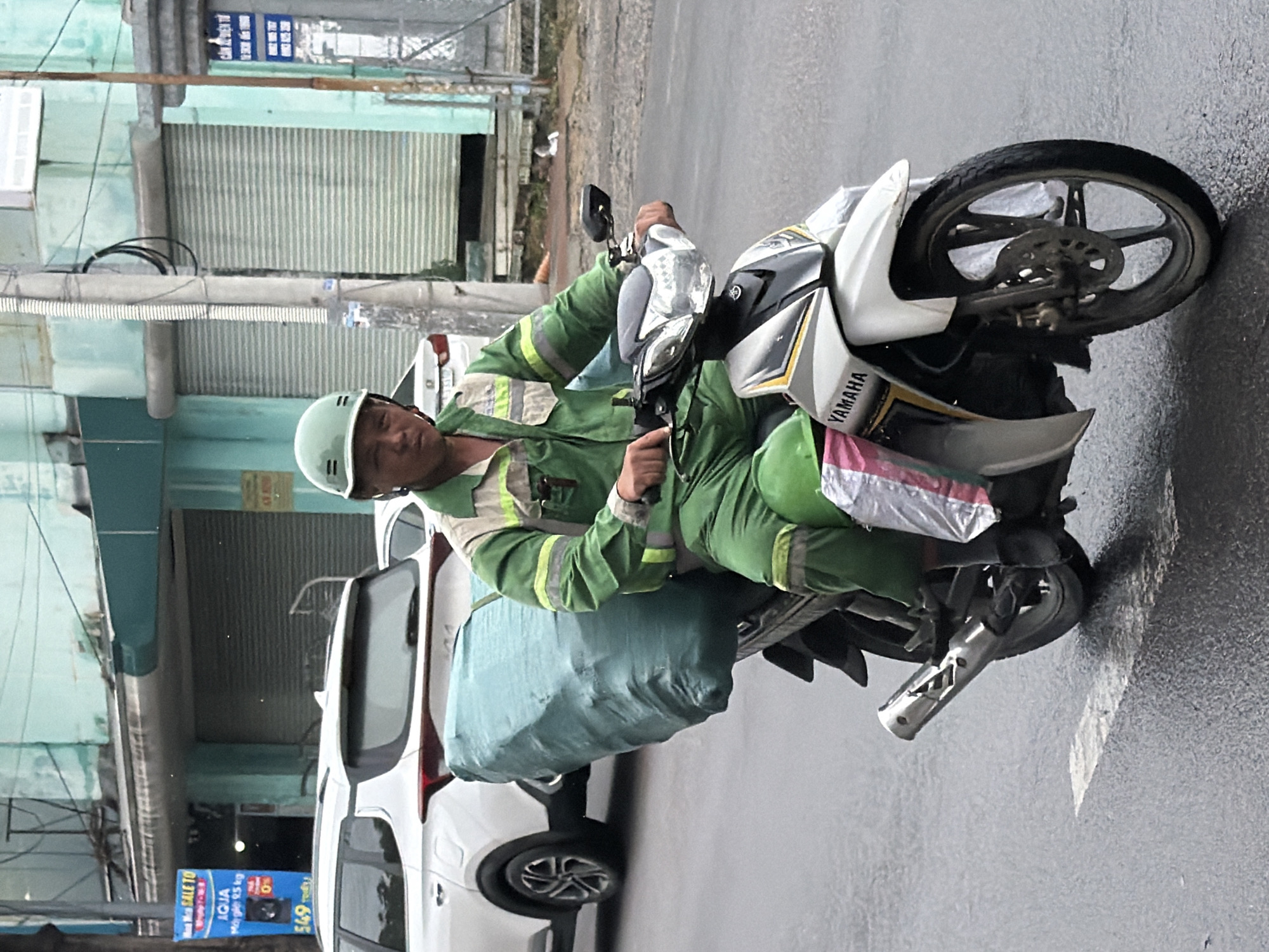 A man wearing a green uniform and helmet rides a white Yamaha motorcycle on a wet asphalt road in Nha Trang, Vietnam. He is transporting a large, teal-colored bag secured to the back of the motorcycle. The man appears to be looking directly at the camera. The background features buildings with roll-up shutters and a white SUV parked on the side of the road. The weather appears overcast and possibly rainy, as indicated by the wet road surface and the man's attire.