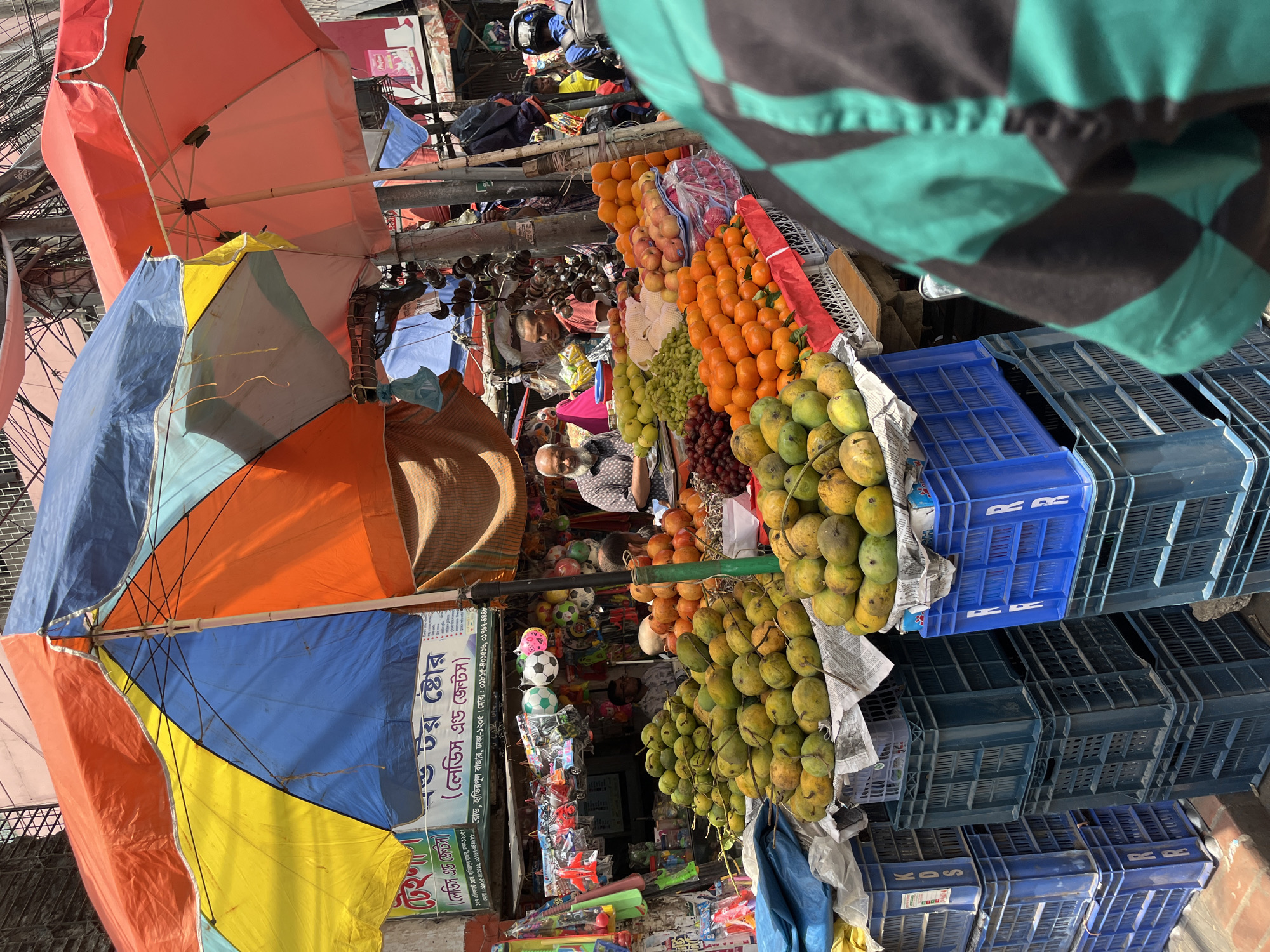The image captures a vibrant street scene in Dhaka, Bangladesh. In the foreground, a colorful array of produce is displayed, including green mangoes, oranges, apples, and grapes. An elderly man with a beard is visible in the center, likely a vendor, surrounded by the fruits. Above the fruit stand, a collection of multicolored umbrellas provides shade, casting a patchwork of colors on the scene. To the left, there is a small shop selling toys, including soccer balls and toy airplanes. The shop's signage includes the words "Ladies and Gents." Stacked blue plastic crates are visible on the right. Overall, the scene depicts a bustling and lively marketplace in Dhaka.