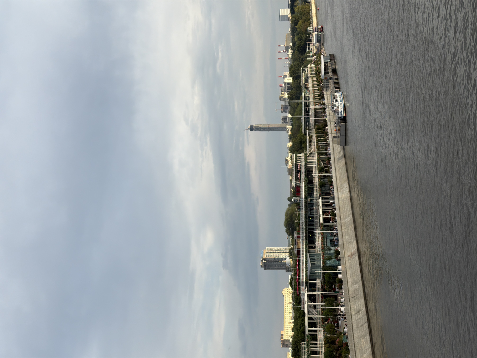 The image depicts a wide river flowing in the foreground, reflecting the overcast sky and the city skyline in the background. Along the riverbank, a long, multi-level structure with white columns and railings is visible, likely a promenade or a building with outdoor seating. People are scattered along this structure, some appearing to be dining or socializing. Several boats are docked along the embankment, including a white passenger boat with blue accents. Further inland, a cluster of buildings forms the city's silhouette, with a tall, slender skyscraper under construction being a prominent feature. Other buildings include a pale yellow long building, a grey high-rise with a sign at the top, and a domed building. In the distance, industrial structures with tall chimneys can be seen. The scene appears to be a public area in Moscow, Russia, likely along the Moskva River.
