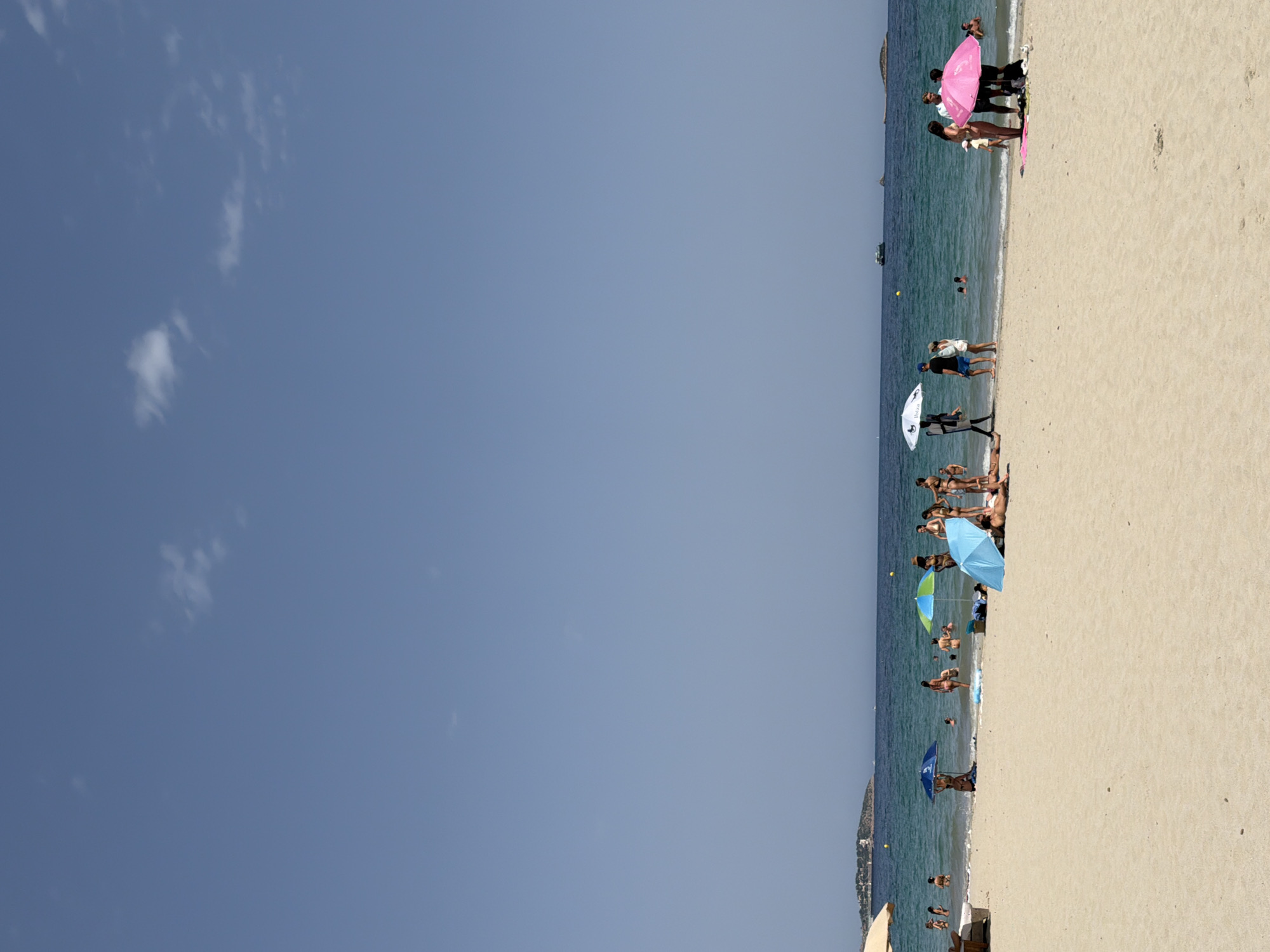 The image depicts a crowded beach scene at Platja D'en Bossa, Spain, under a clear blue sky with scattered white clouds. The foreground is dominated by a vast expanse of light-colored sand. In the middle ground, the turquoise water of the Mediterranean Sea is visible, with numerous people swimming, wading, and standing in the shallow areas. Beach umbrellas of various colors, including a large beige one on the left, a blue one, a blue and green striped one, and a pink one on the right, provide shade for beachgoers. Several individuals are seen lounging on the sand, some appearing to sunbathe. Groups of people are standing and interacting, some under umbrellas, while others are in the water. In the background, a low-lying island with buildings and vegetation is visible on the horizon. Several boats are also present on the sea. The overall atmosphere is one of a bustling summer beach day.