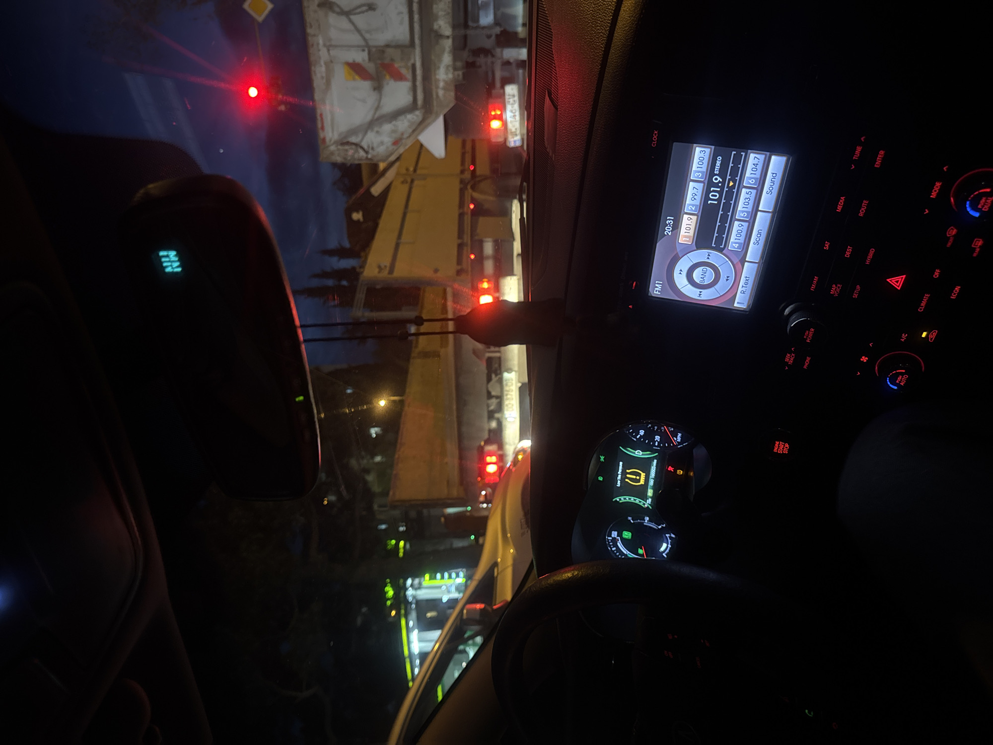 The image captures a nighttime scene from the interior of a car, looking out through the windshield onto a street in Tbilisi, Georgia. The car appears to be stationary, possibly in traffic or at a red light, behind a large commercial vehicle, likely a garbage truck or a similar utility truck, identifiable by its bulky structure and multiple red taillights.
Inside the car, the illuminated dashboard and infotainment system are prominent. The infotainment screen displays "20:31" as the time and shows the radio tuned to "FM1 101.9 STEREO," with other selectable frequencies such as 99.7, 100.3, 100.9, 103.5, and 104.7. Surrounding the screen are various buttons for car functions like "MEDIA," "ROUTE," "HYBRID," "CLIMATE" controls (A/C, OFF, ECON), "MODE," "PUSH DUAL," "PUSH POWER," "SEEK TRACK," and "PHONE." The instrument cluster is partially visible on the right, displaying a "Low Tire Pressure" warning symbol, "0 MPH," and "165503" which likely indicates the vehicle's mileage in kilometers. A digital compass in the rearview mirror displays "NW."
Outside the car, the large truck in front has a visible license plate that reads "AY 493 CY." Beyond it, other red lights suggest more vehicles or traffic signals further down the street. The ambient light from street lamps and other distant buildings reflects on the wet road or windshield, creating glints. The overall setting is dark, emphasizing the artificial lights of the vehicles and the car's interior.