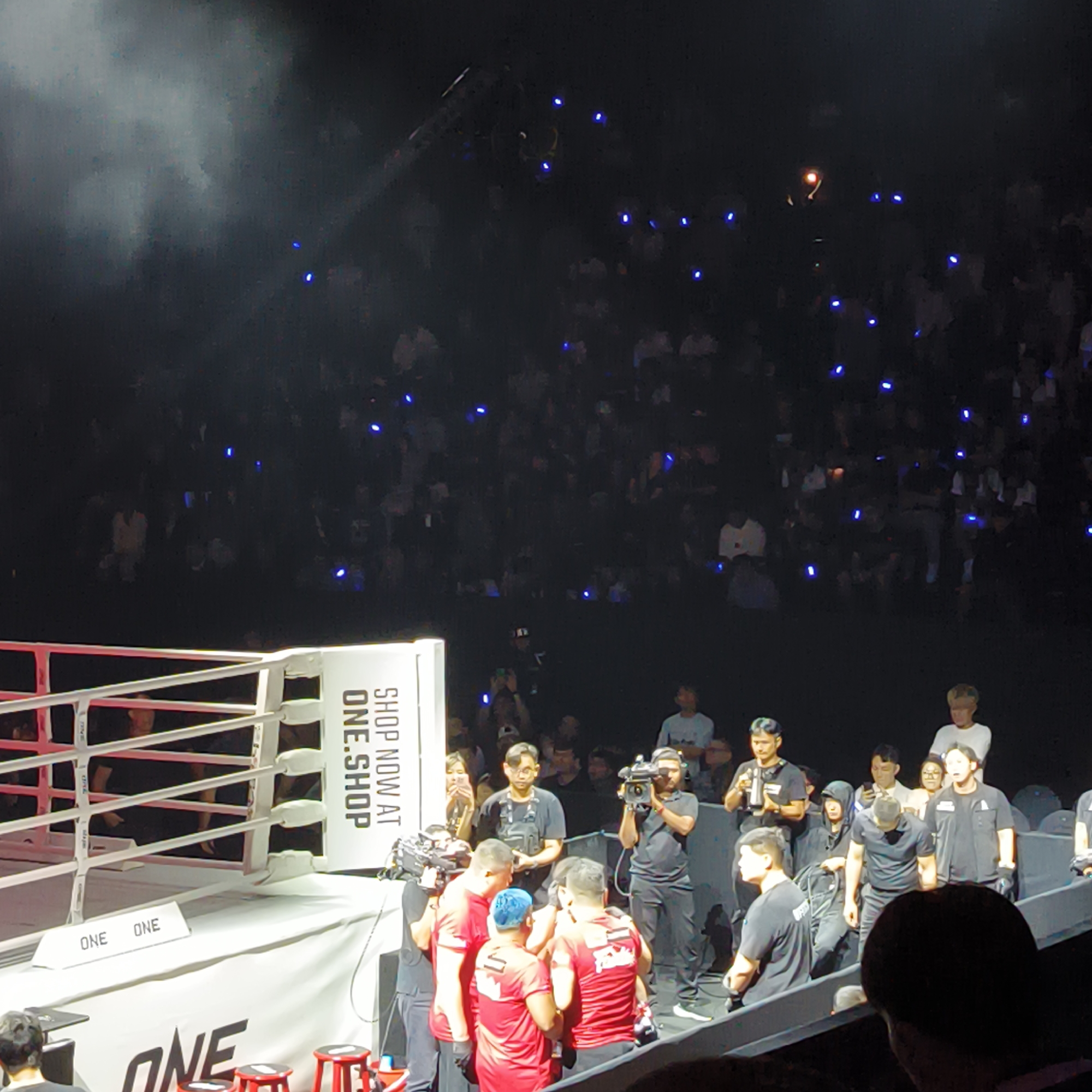 The image depicts a ONE Championship mixed martial arts event, likely in Bangkok, Thailand. An elevated boxing ring dominates the foreground, with the ONE Championship logo prominently displayed. Red and white rope is visible.
A large crowd fills the background, with many spectators holding up small blue lights or phones, creating a speckled effect. In the midground, a group of people are gathered near the ring. A camera crew films them. Several men wear red shirts with a logo, potentially coaches or team members. A man with bright blue hair is also visible. Other people in the area appear to be event staff, wearing black clothing. The environment appears to be a large, indoor arena, illuminated with spotlights.