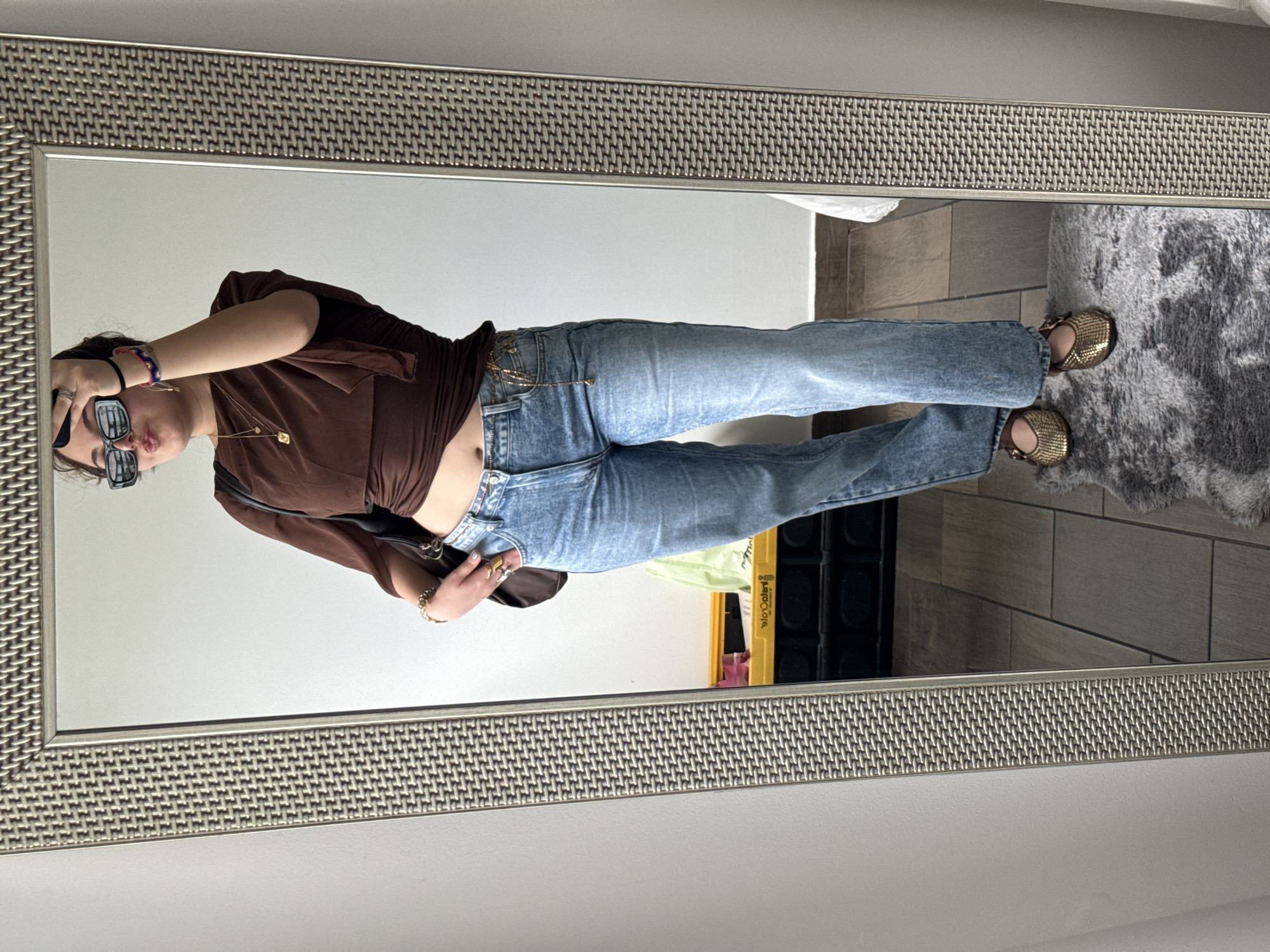 A woman takes a mirror selfie. She wears blue jeans and a brown blouse. She is standing in a room with light-colored walls. A grey rug is visible in the reflection. A yellow and black object is also visible behind her. It is not possible to determine the exact location in Mexico City from this image.