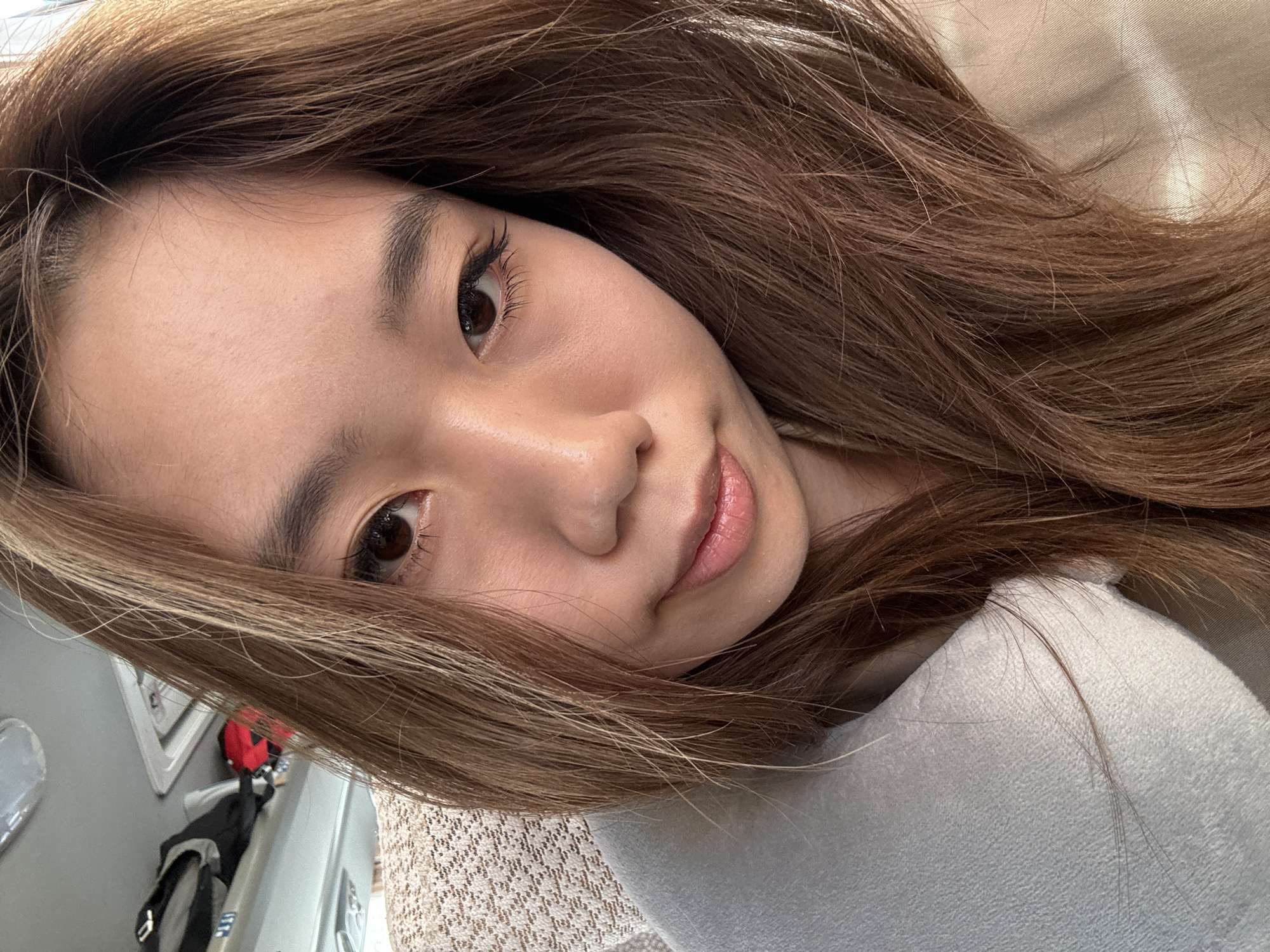 A close-up, portrait-style photograph shows an Asian woman with long, brown hair. Her skin is light-toned. Her eyes are brown. She has dark eyebrows and long eyelashes. Her lips are full and appear to have a glossy coating. She is wearing a light-colored, possibly white or gray, soft-looking garment. The background is blurred. A partial view of what appears to be a wall or surface with a textured beige pattern and a gray surface is visible. The photo is taken at an angle. There is no verifiable information to determine this photograph was taken in Wuwei, China.