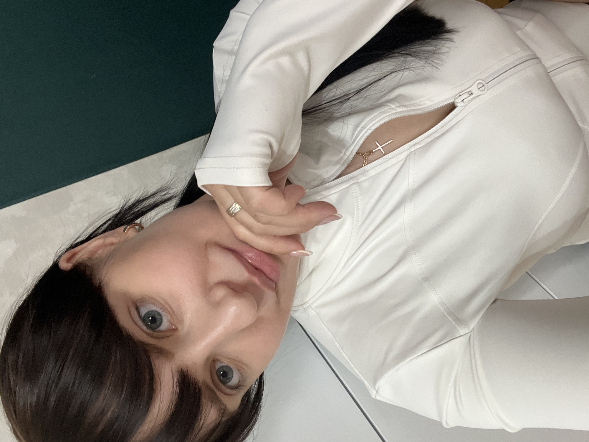 A rotated, close-up portrait depicts a young woman with dark hair and wide blue eyes looking directly at the viewer. Her dark hair is styled with bangs, framing her face. She is wearing a white, long-sleeved top that features a prominent zipper on the chest, partially undone to reveal a small golden cross necklace. Her right hand, adorned with a silver-toned ring on the ring finger and manicured nails with white tips, is gently placed near her chin and lower lip. The background features a dark green wall on the left side, meeting a lighter, possibly white or light grey, surface or floor on the right and bottom, suggesting a simple indoor setting. The woman appears to be posing for the camera with a calm and engaged expression. There is no visible text in the image. The provided location context is Марковский, Russia.