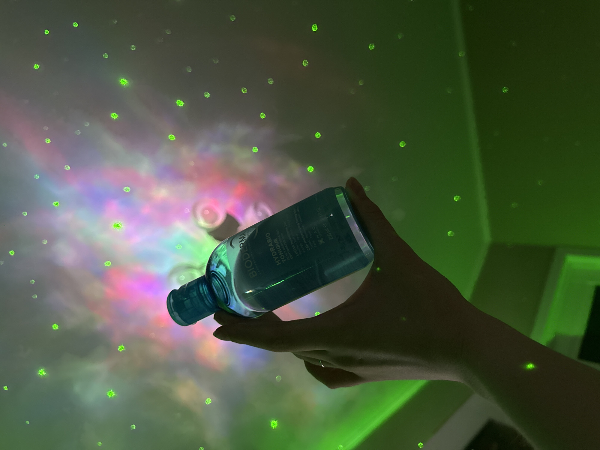A hand is holding a bottle of BIODERMA HYDRABIO TONIQUE in an indoor environment. The background is illuminated with a combination of green dots and a colorful light pattern that includes pink, purple, blue, and white. The lights appear to be projected onto a wall or ceiling. The hand holding the bottle is partially silhouetted against the light.