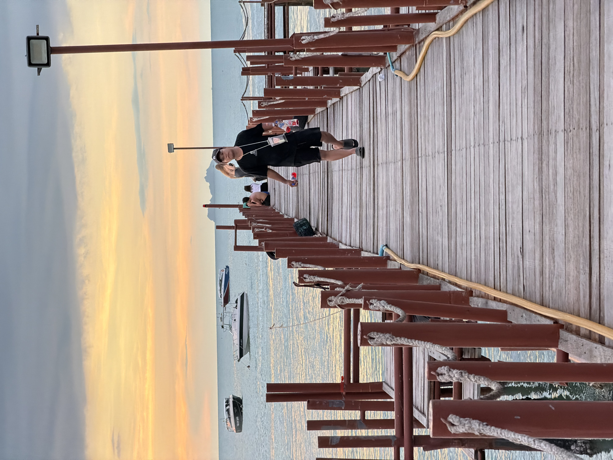 This is a medium shot image taken on a wooden pier in Baan Bang Rak, Thailand. The sky is a blend of gray and orange clouds, indicative of sunset. Several boats are visible on the calm sea in the background. In the foreground, a man wearing a black t-shirt, black shorts, a black baseball cap, and dark sandals is walking towards the camera. He is carrying a water bottle in his left hand and a clear pouch around his neck. To his right, a yellow hose runs along the edge of the pier. Other people are seen sitting and standing further down the pier in the background. The pier is constructed of wooden planks and has red-colored railings with ropes attached.