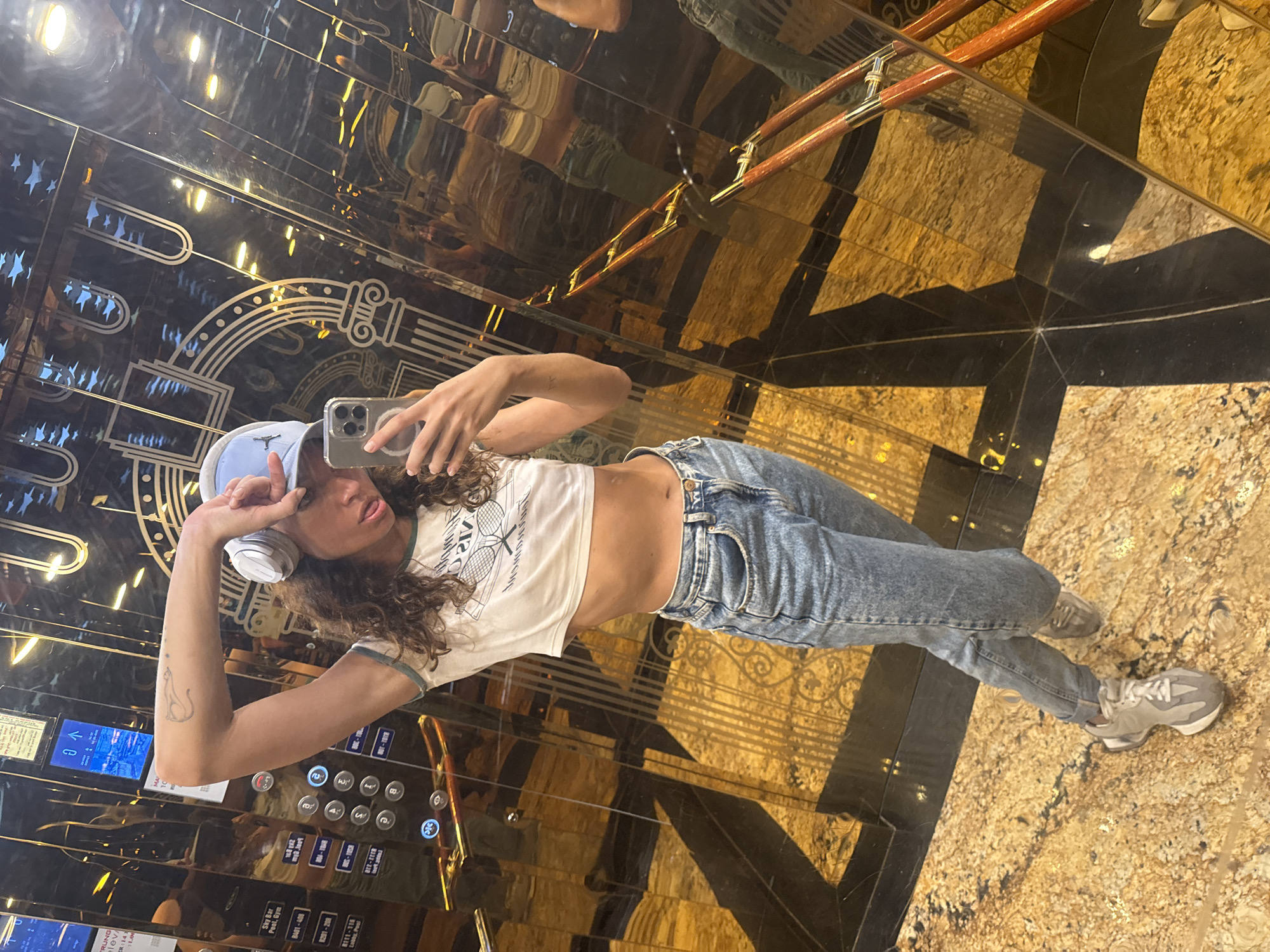 A young woman in jeans, a white crop top, and a light blue baseball cap is taking a selfie in an elevator. She has headphones around her neck. The elevator has a mirrored interior with decorative inlays. A control panel with buttons and digital displays is visible on the left side of the frame. A handrail is reflected in the top right corner. The elevator floor is marble with a starburst pattern. Based on some clues in the image such as the panel saying "Sky Bar, Pool, Gym" it is possibly taken in a hotel elevator. The writing on the elevator display shows possible location as Da Nang, Vietnam.