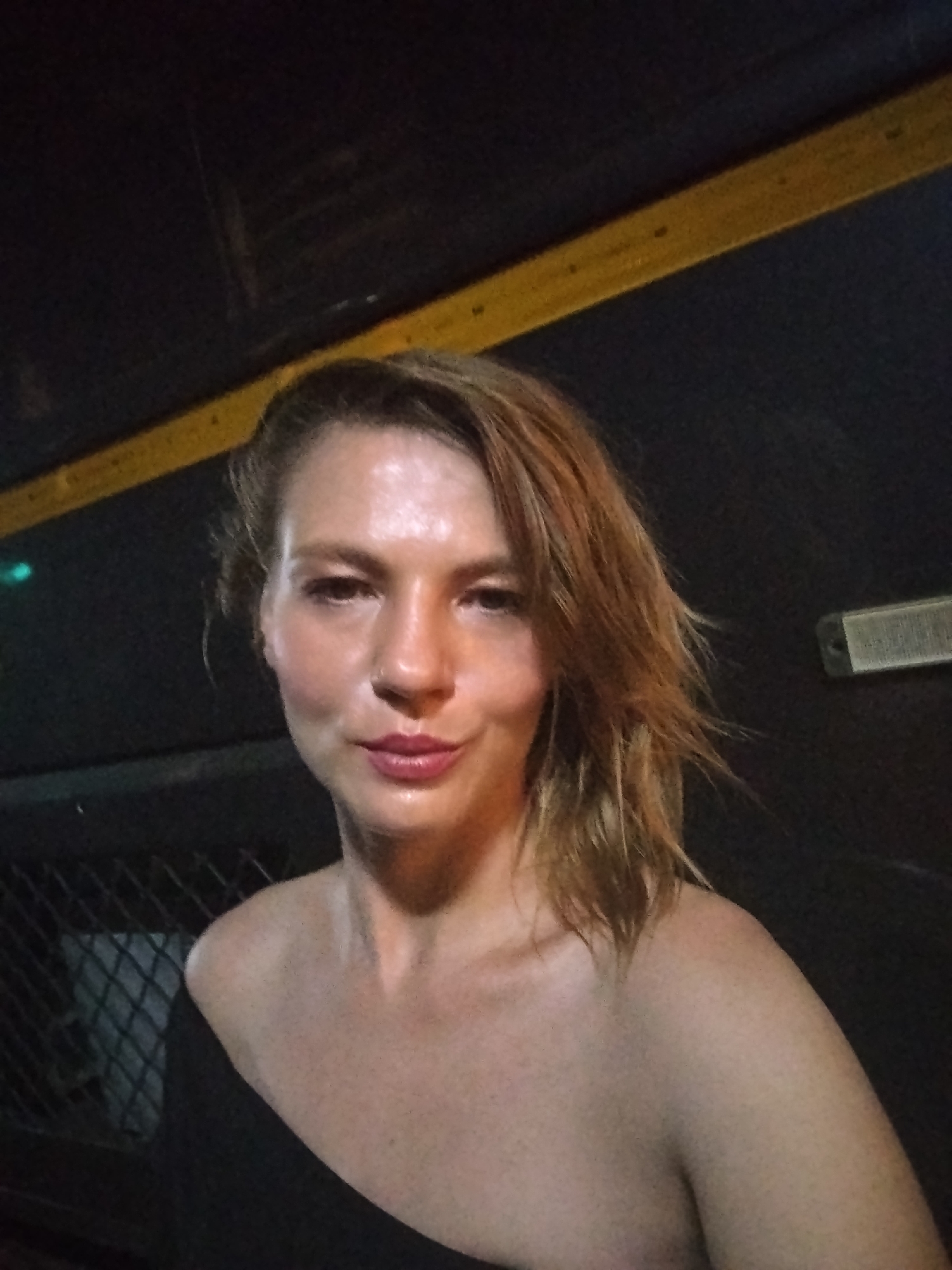 The media file presents a close-up portrait of a woman, likely a selfie, set in what appears to be a dimly lit indoor or covered environment in Kalamassery, India. The woman is the central subject, seen from the mid-chest up, looking directly at the camera with a calm, neutral expression. She has light skin, and there is a noticeable sheen on her forehead, nose, and shoulders, which could suggest sweat or a humid atmosphere. Her medium-length reddish-brown hair is styled to one side, covering part of her left shoulder. She is wearing a dark, off-the-shoulder top or dress, revealing her collarbone and shoulders. A subtle nose piercing is visible on her right nostril, and her lips are painted with light pink lipstick.
The background is predominantly dark. A prominent horizontal yellow stripe or bar runs diagonally across the upper part of the frame, possibly indicating a structural beam, handrail, or part of a vehicle interior. To the left, a faint green light source is visible. On the right, a rectangular object with a textured, possibly metallic, surface is discernable, which could be a vent or a light fixture. In the lower-left background, a dark, diamond-patterned mesh or fence is partially visible. The overall setting appears utilitarian, possibly an urban public space or a form of transport, consistent with the location context of Kalamassery, India, during nighttime or in low light conditions. No visible text is present in the image.