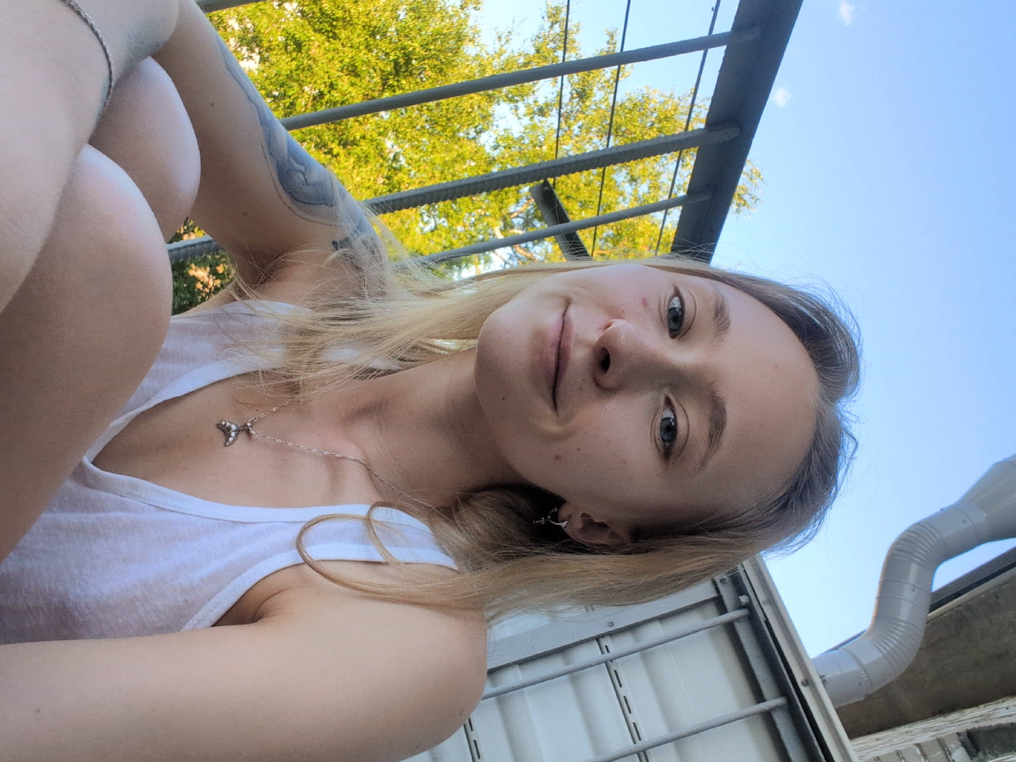 The image depicts a young Caucasian woman with blonde hair and blue eyes, wearing a white tank top and a silver necklace. She has a tattoo on her left arm and an earring in her right ear. The woman is seated outdoors, with a railing and trees visible in the background, along with a downspout and a building facade. The sky is clear and blue. The scene appears to be a balcony or porch area. There are no explicit indicators of the city of Veliky Novgorod, Russia.