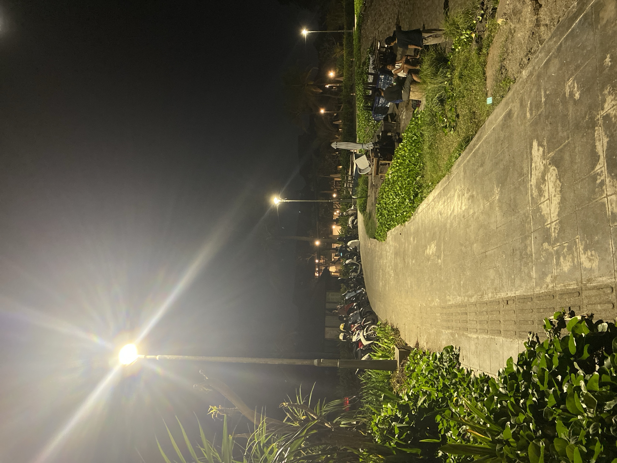 The image depicts a nighttime scene in North Kuta, Indonesia. A concrete path, illuminated by streetlights, curves through the frame. To the right of the path, vegetation lines the edge. In the distance, people are seated at tables. A row of parked scooters and motorcycles extends along the path. Streetlights are visible at intervals along the path, with one streetlight dominating the lower left of the frame.
