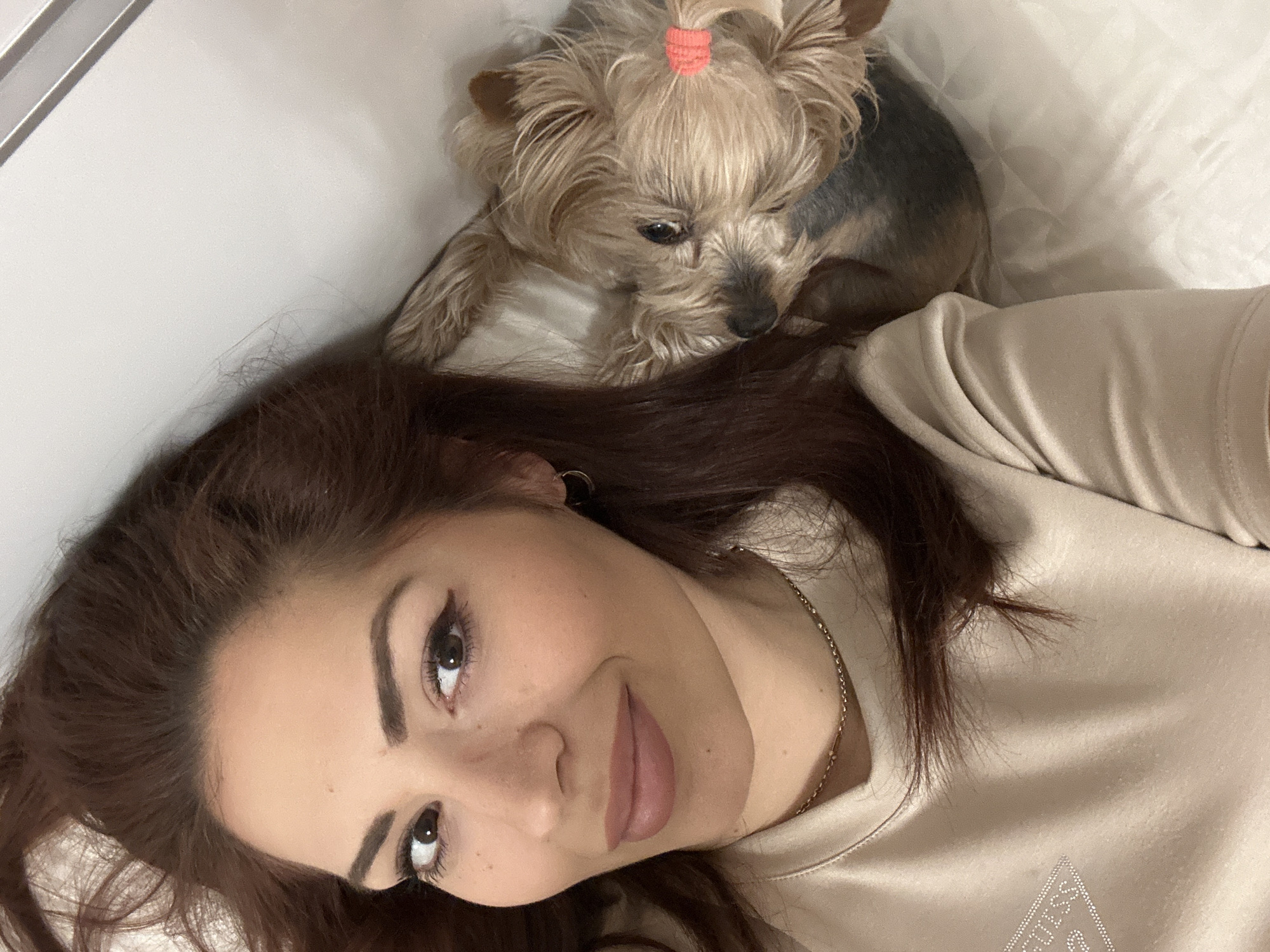 A woman is lying on a white quilted surface, possibly a bed. She has long brown hair, fair skin, and is wearing a tan top. Around her neck is a thin gold chain necklace. A small brown and gray dog with a pink hair tie is sitting on her head/shoulder area. The background appears to be a white wall. There is no information in the image to indicate this photo was taken in Sofia, Bulgaria.