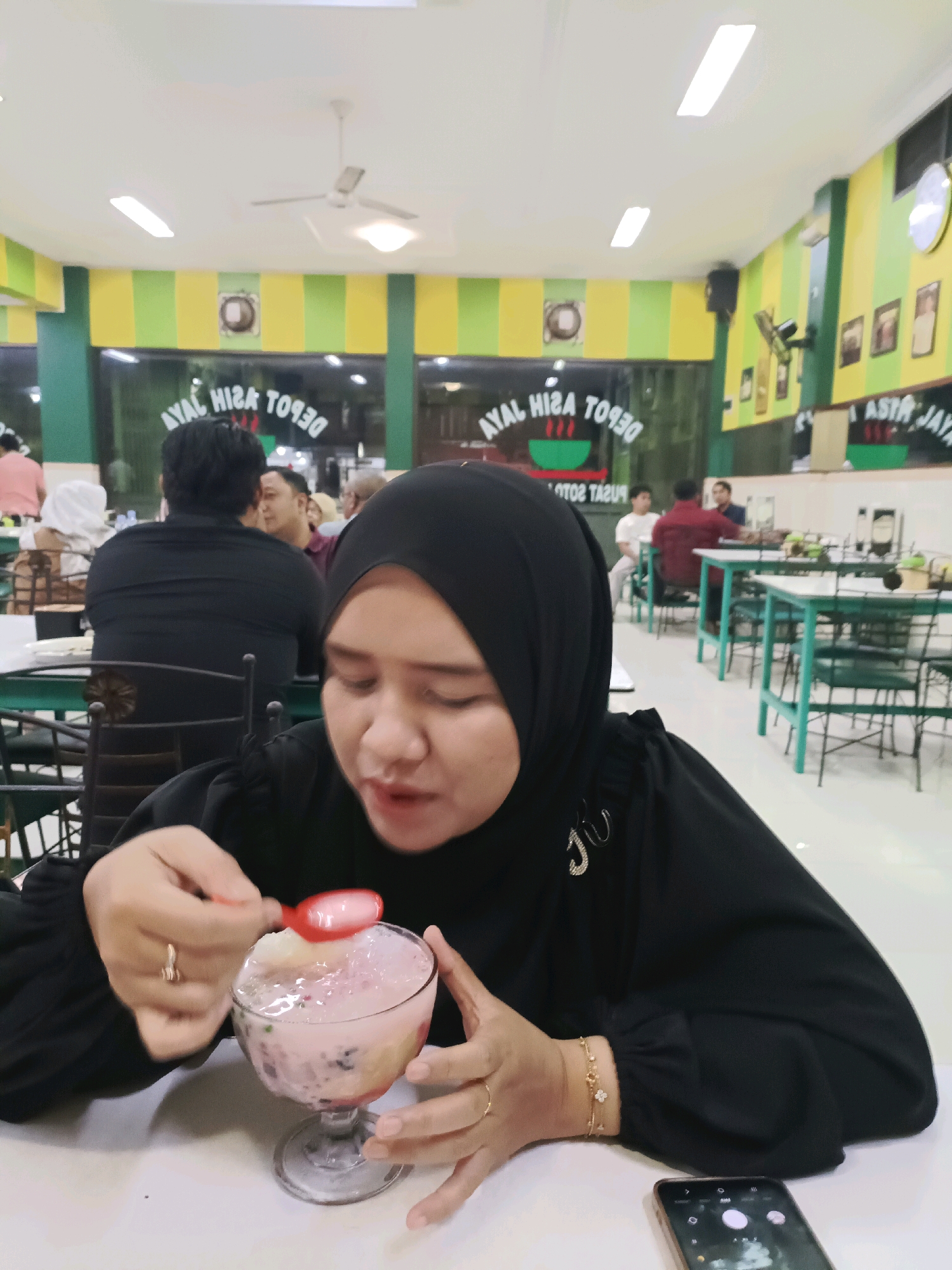 A woman wearing a black hijab and black blouse is sitting at a white table in a restaurant, eating a pink dessert with a red spoon. She has a ring on her left ring finger and a bracelet with a small flower charm on her right wrist. To her right, on the table, is a smartphone displaying a camera interface. The restaurant has green and yellow striped walls, and tables with green chairs are visible in the background. Signs with text that appears to be in Indonesian are displayed on the wall, including "AYAL HISA DEPOT" and "ASTRA JAYA". The overall ambiance suggests a casual dining establishment in Dapur, Indonesia.