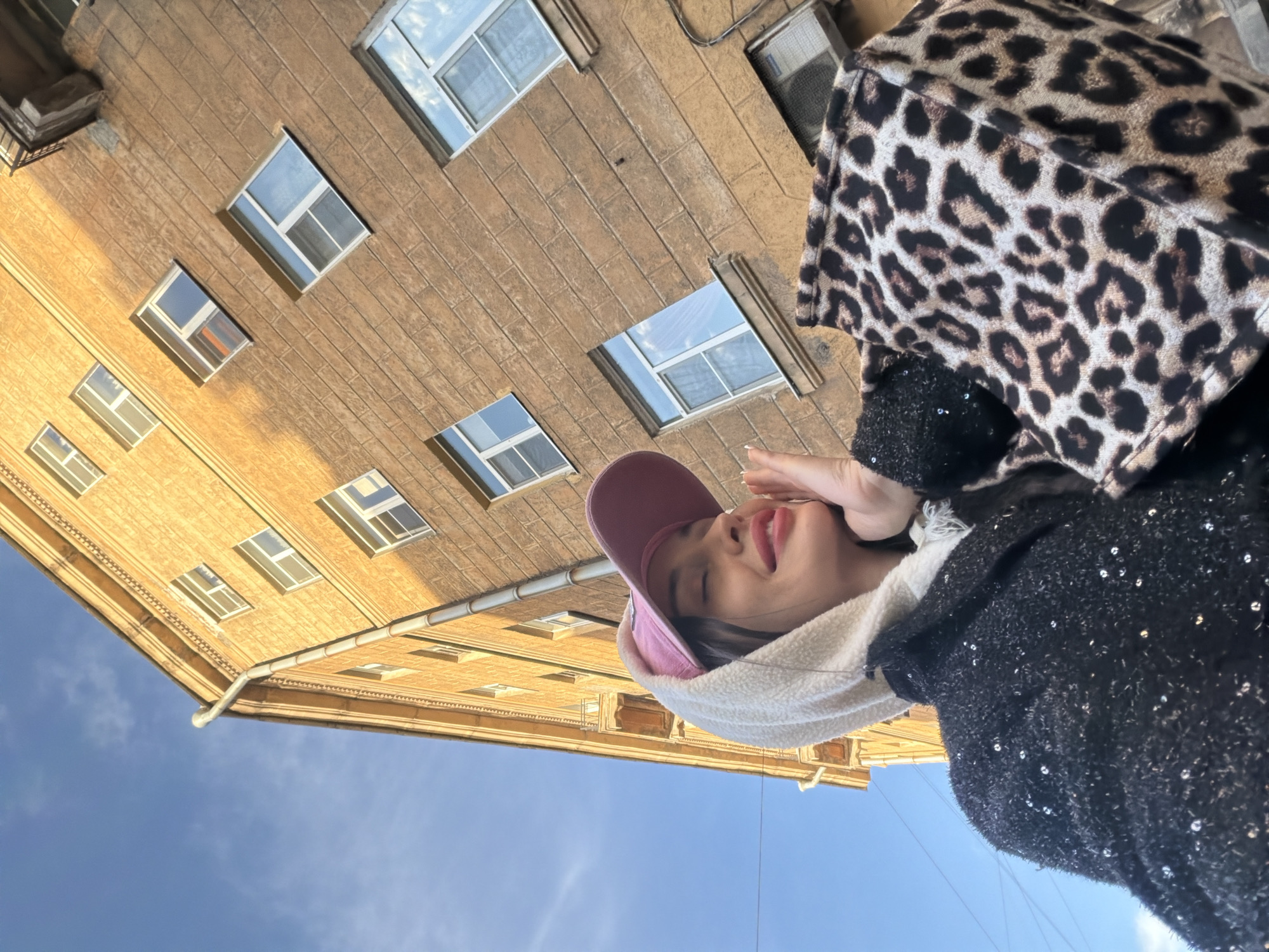 A low-angle, rotated outdoor shot of a woman standing in Saint Petersburg, Russia, in front of a brown, multi-story building with symmetrical rows of rectangular windows. The windows reflect the sky, suggesting they are glass. The sky is clear blue. The woman is wearing a pink cap, a white hooded sweatshirt, and a dark, sequined jacket with a leopard-print bag over her shoulder. She is holding her hand up to her face, appearing to smile. The orientation of the photograph suggests the building looms over the person. The photo is well-lit with natural sunlight.
