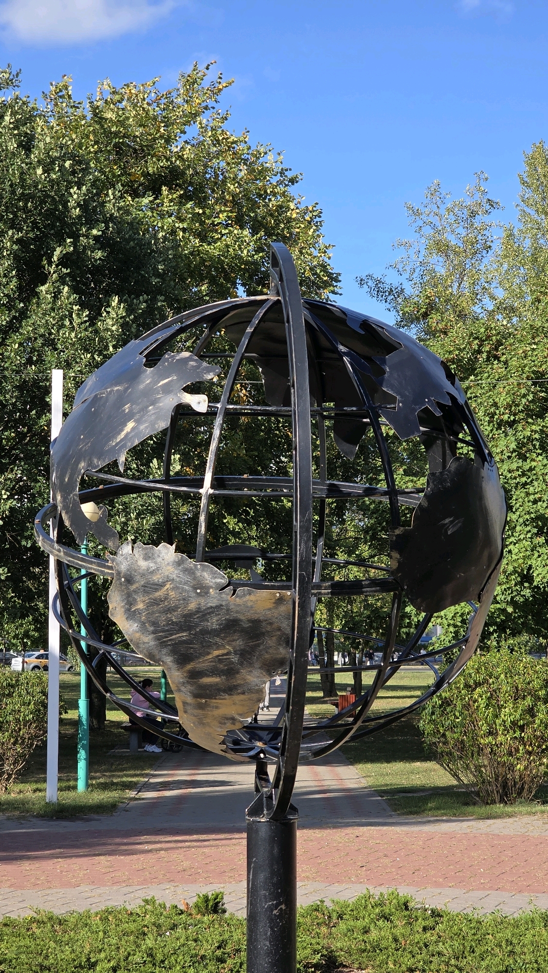 A metal sculpture of a globe is situated in an outdoor park-like setting. The globe is supported by a black, cylindrical metal pole. The globe itself is constructed from black metal strips that form a spherical frame, with cutouts representing continents. The continents are depicted in a rough, metallic finish, appearing somewhat worn and textured.
The background features lush green trees with some yellowing leaves, suggesting it may be autumn. The sky is a clear, bright blue with a few wispy clouds. In the mid-ground, there is a paved walkway made of red and grey interlocking bricks. A person in a pink shirt is seated on a bench to the left of the walkway, partially obscured by bushes. Several other people are visible further in the background, walking along the path or sitting on benches. To the far left, a white pole and a green pole are visible.
The overall scene conveys a sense of a public outdoor space, possibly a park or square, in a city environment. The globe sculpture serves as a prominent focal point. This sculpture is located in the city of Brest, Belarus, specifically on Sovetskaya Street, which is a pedestrian street known for its art objects and historical buildings.