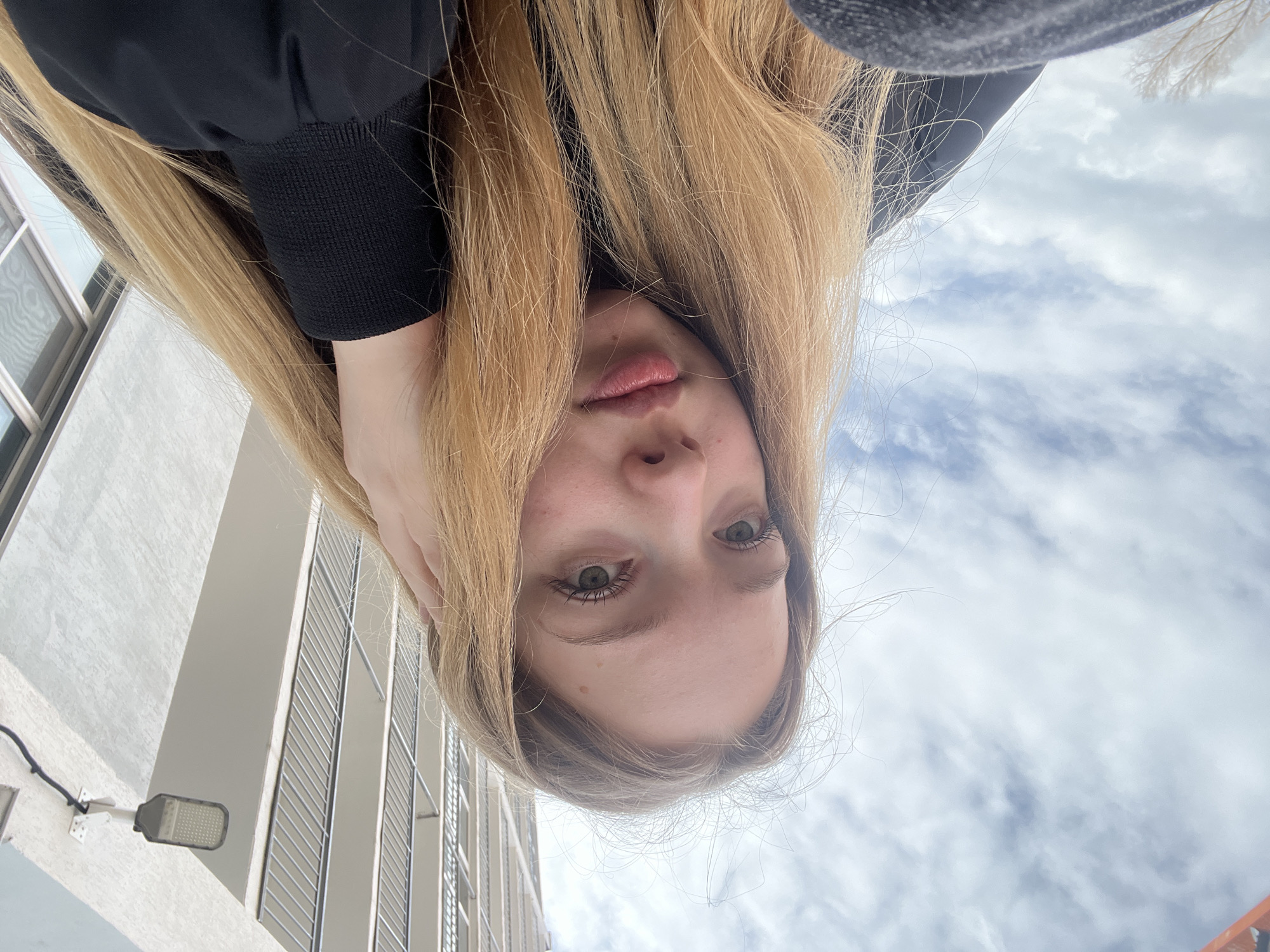 A person is upside down taking a selfie. The person has light blonde hair, fair skin, and light-colored eyes. They are wearing a dark-colored jacket or sweater. The background includes a building with multiple windows and metal railings, a street light, and a cloudy sky. The location may be the city of Chaikovsky, Russia.