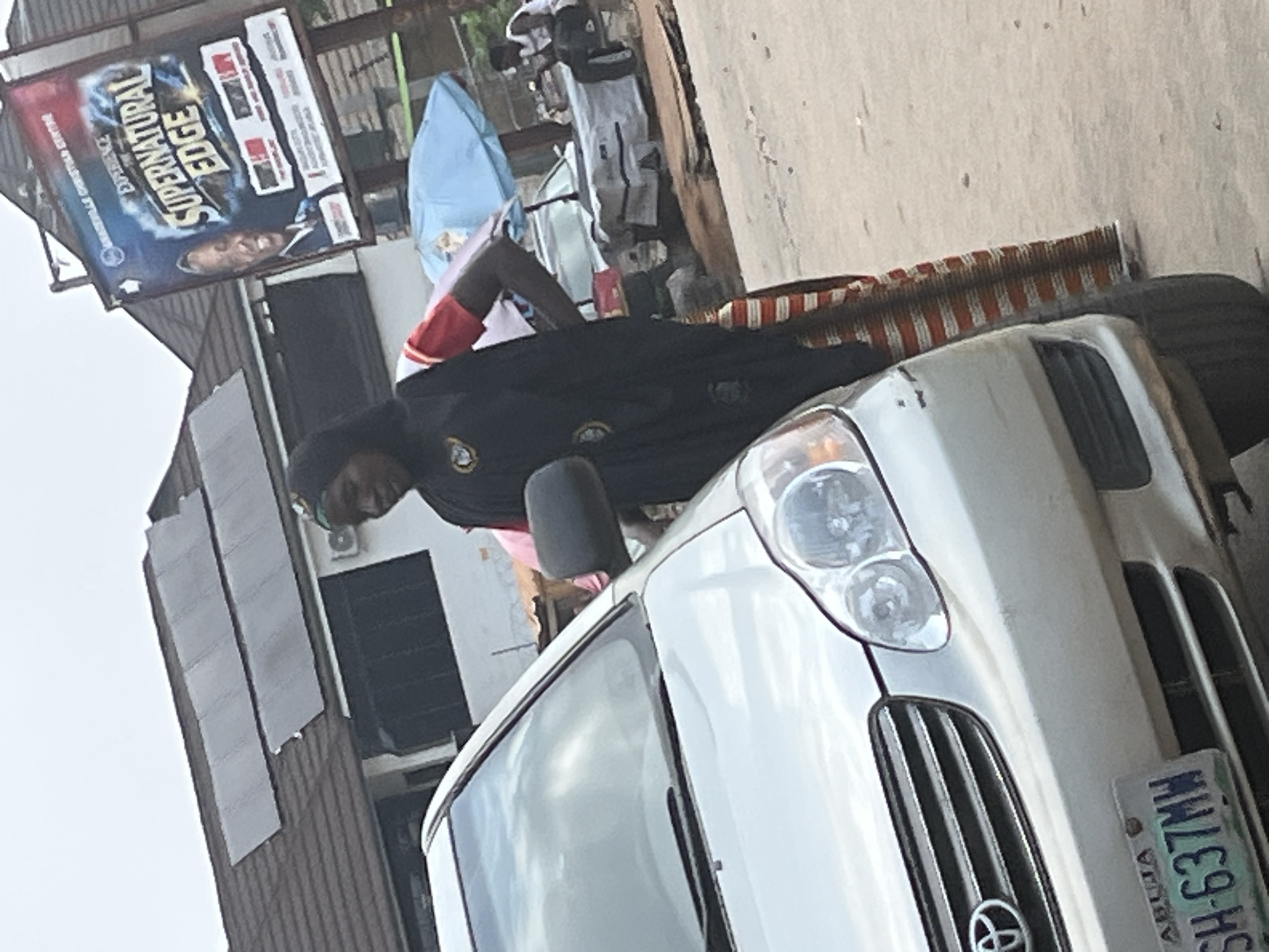 In a street scene in Abuja, Nigeria, a silver Toyota Corolla is parked. In the foreground, the front passenger side of the car is visible, including its license plate which reads "ABUJA SH-637MW". To the right of the car, a woman wearing a black hijab and a red shirt stands with her hand on her hip. She is wearing a striped skirt. Behind her, a billboard advertises "EXPERIENCE THE SUPERNATURAL EDGE" for a Christian center. Further in the background, a man sits on a bench near a stall with a blue umbrella, and another person is partially visible behind him. The environment appears to be an outdoor, urban setting with buildings and a cloudy sky.