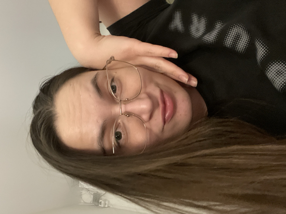 A woman with long brown hair and glasses lies on her side. She wears a black t-shirt with a white design. Her hand rests near her face, supporting her head. She looks directly at the camera with a gentle smile. The background is a plain, light-colored wall. The image is slightly rotated.