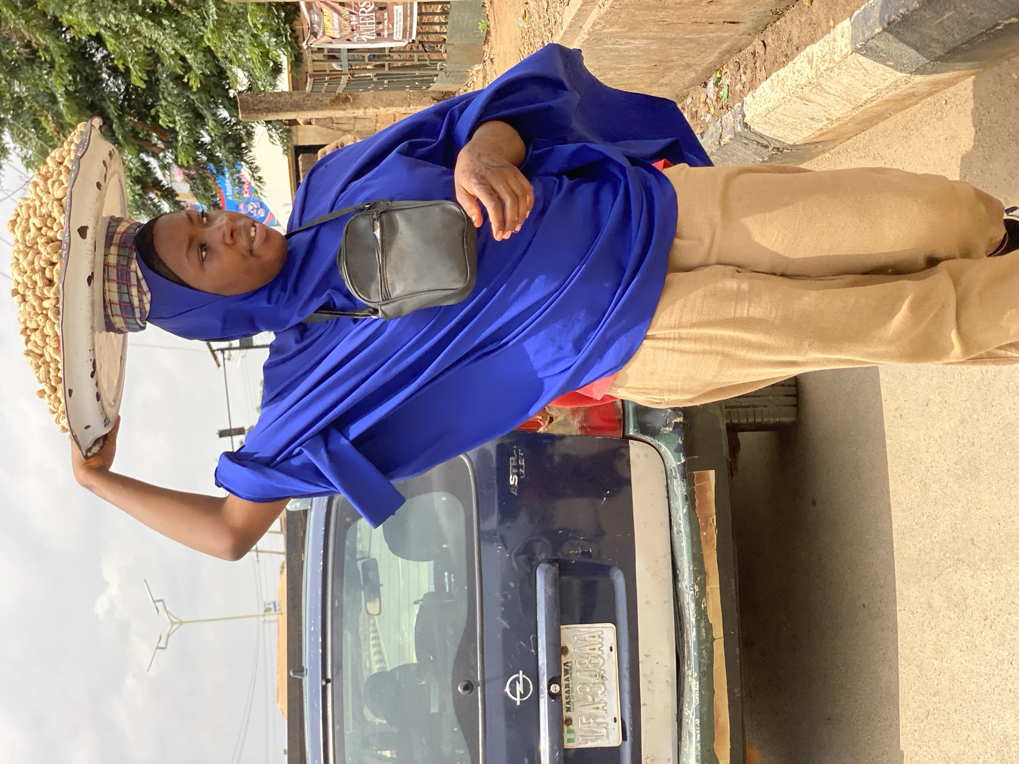 A female vendor stands in an outdoor street setting, likely engaged in hawking, in Kafanchan B, Nigeria. She is wearing a royal blue hijab and a matching loose-fitting blue garment over light brown trousers. A small black shoulder bag is worn across her body. On her head, she expertly balances a large, white, oval-shaped tray overflowing with shelled peanuts. Her right arm is bent, possibly steadying the tray. She looks slightly upwards and to her right with a calm expression and a faint smile.
Behind her, partially visible, is the rear of a dark blue vehicle, which appears to be an Opel, featuring its distinctive logo and a Nigerian license plate. The license plate reads "NASARAWA LFA-34BAA". The background includes green foliage from trees, a street curb, and a bright, slightly cloudy sky, indicating daytime. Some distant, partially legible commercial posters are visible in the upper right background, including fragments like "CRON..." and "A taste...". The lighting suggests it is sunny, with soft shadows. Faint patterns, possibly henna, are visible on her right hand.
