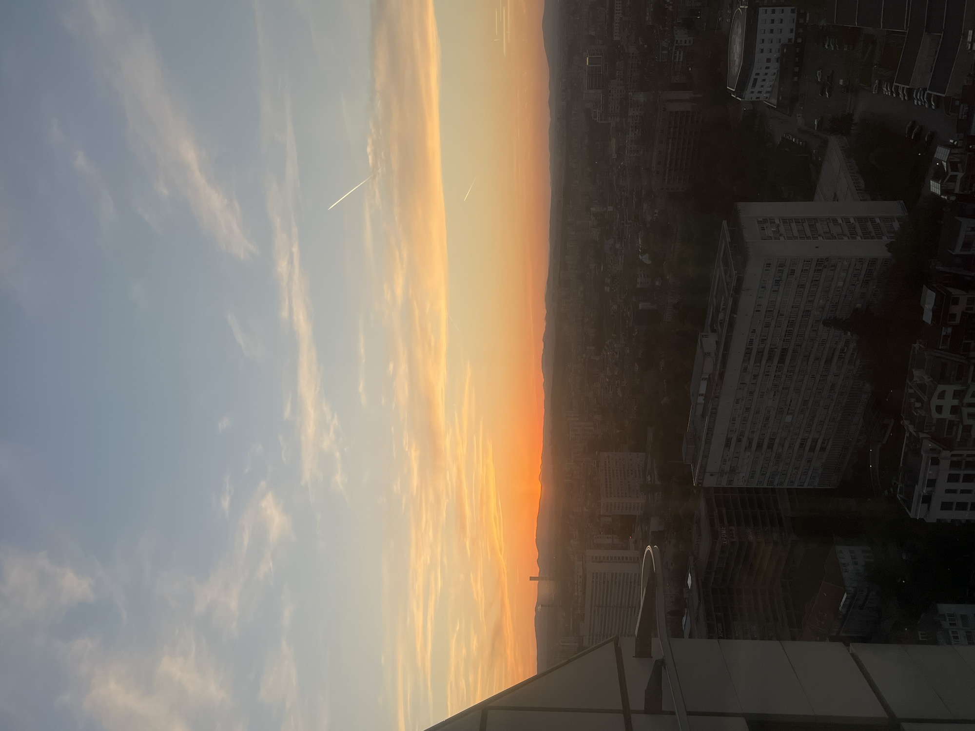 The image displays a panoramic view of a city and sky, likely captured during either sunrise or sunset from a high vantage point, such as a rooftop. The scene is horizontally divided, with the sky occupying the upper part and the cityscape the lower.
The sky transitions from a soft, light blue at the top to warm hues of orange, yellow, and subtle pink near the horizon, indicating the presence of the low sun. Wispy, elongated clouds are scattered across the blue portion of the sky, catching the ambient light. Two prominent white contrails, left by aircraft, are visible, one clearly defined in the blue sky and another fainter one closer to the colorful horizon.
Below the horizon, the sprawling cityscape stretches into the distance, characterized by numerous multi-story buildings, many appearing as dark silhouettes against the brighter sky due to the low sun angle. The urban landscape gives way to a range of mountains or hills on the distant horizon, which are also silhouetted. In the immediate foreground, on the bottom right, a portion of a dark, angular rooftop or building structure is visible, confirming the elevated perspective.
The time of day is clearly either early morning or late evening, marked by the dramatic sky colors and long shadows. The weather appears clear with minimal cloud cover, allowing for excellent visibility of the distant landscape. While no specific landmarks are identifiable, the combination of urban development and a mountain backdrop is consistent with the context of Sofia, Bulgaria, possibly showing the Vitosha mountain range. The scene evokes a sense of tranquility and the grandeur of an urban setting at a transitional time of day. No visible text is discernible in the image.