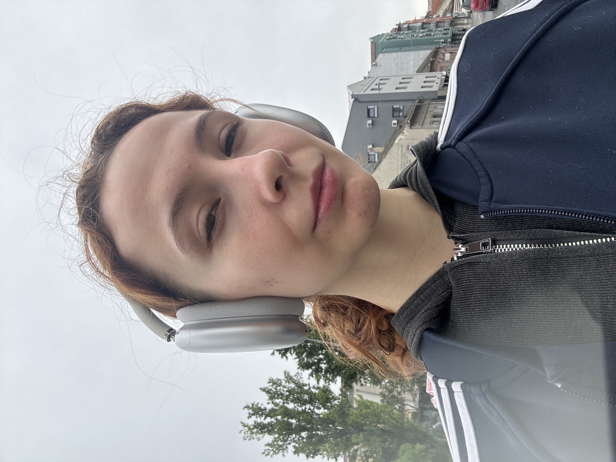 A vertical self-portrait features a young person with light brown or auburn hair looking directly at the camera with a neutral, slightly relaxed expression. They have fair skin and wear large, silver-grey over-ear headphones, possibly Apple AirPods Max.
The person is dressed in a dark blue athletic jacket with prominent white stripes running down the sleeves, indicative of the Adidas brand, whose logo is partially visible. Beneath the jacket, a black zippered top is worn.
The background depicts an urban environment under an overcast, light grey sky. Multi-story buildings with various grey and white facades are visible behind the person, with one structure in the upper right featuring red details on its roof. Below the buildings, several green trees indicate a street or park setting. The overall impression is that of an outdoor urban scene during daytime, likely a cloudy day. Given the context, the location is Novi Sad, Serbia. The person appears to be casually enjoying music or a podcast while outdoors.