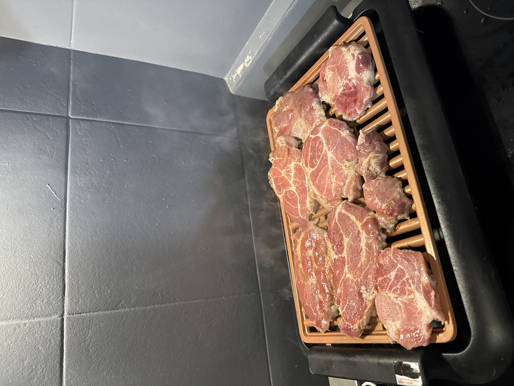 An overhead, slightly angled shot captures several pieces of raw, seasoned meat cooking on an electric indoor grill in a kitchen setting in Stara Zagora, Bulgaria. The grill features a distinctive copper or bronze-colored non-stick grate, holding approximately ten individual cuts of what appear to be pork steaks or chops. The meat is reddish-pink with visible marbling and white fat, lightly coated with seasonings. Wisps of steam are rising from the cooking surface, indicating the grill is actively heating.
The electric grill itself has a dark grey or black exterior, with a prominent handle and base. A small, partially obscured label with illegible text and a small icon is visible on the front right base of the grill, likely a warning or instruction sticker.
The background consists of dark grey or black square tiles, which appear textured and form a countertop or wall surface. These tiles extend to the left and rear of the grill, with distinct grout lines. Above the darker tiles, there is a transition to a lighter grey or off-white wall or cabinet, showing some minor wear or debris where the two surfaces meet. There are no people visible in the image, and the time of day and weather are not discernible from this indoor scene.