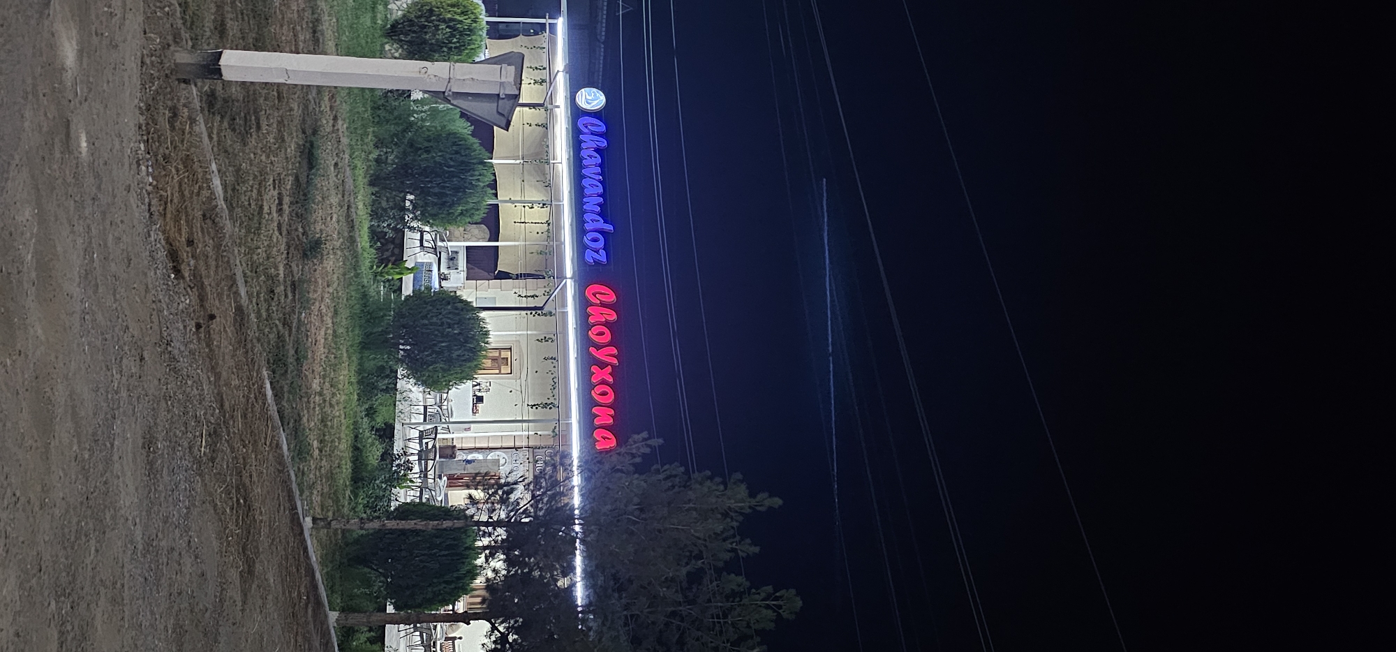 The image depicts a nighttime scene in Sheyhancha, Uzbekistan, featuring a roadside establishment with a prominent neon sign. The sign reads "Chavandoz Choyxona" in alternating blue and red letters. The establishment appears to be a cafe or restaurant, with outdoor seating visible under a covered area. Several small trees and bushes are planted around the perimeter of the building. A concrete post is located in the foreground, to the left of the building. The sky above is dark, indicating it is nighttime, and there are visible power lines stretched across the upper portion of the frame. The overall impression is of a commercial establishment situated along a road in a rural or semi-rural setting.