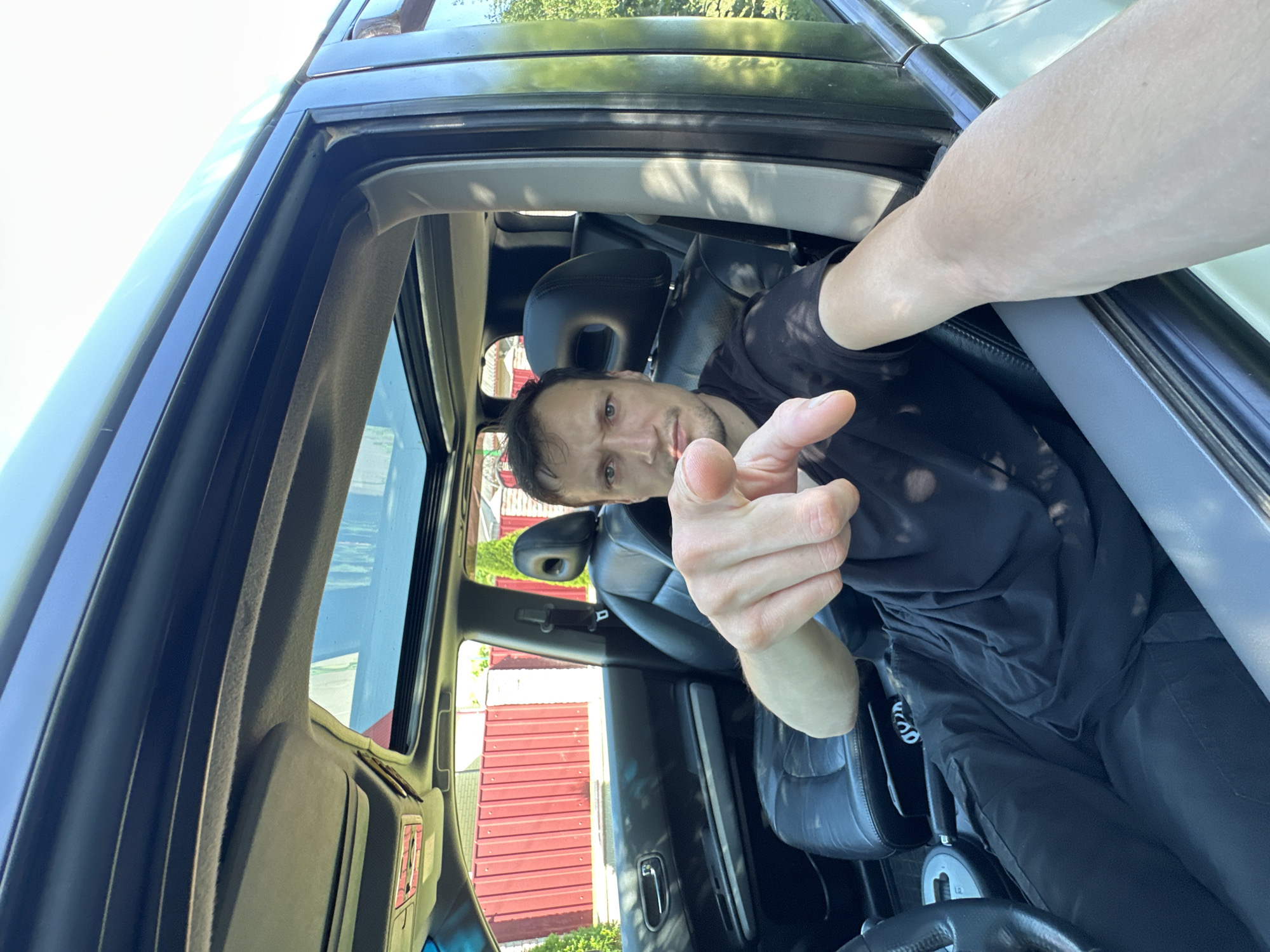 A man is sitting in the driver's seat of a car, looking directly at the camera and pointing his index finger towards it. The car's interior is visible, including black leather seats, a sunroof, and the dashboard. The man is wearing a black t-shirt and black pants. The car is parked outdoors, with a red fence and greenery visible in the background. The scene appears to be in Shatava, Ukraine.
