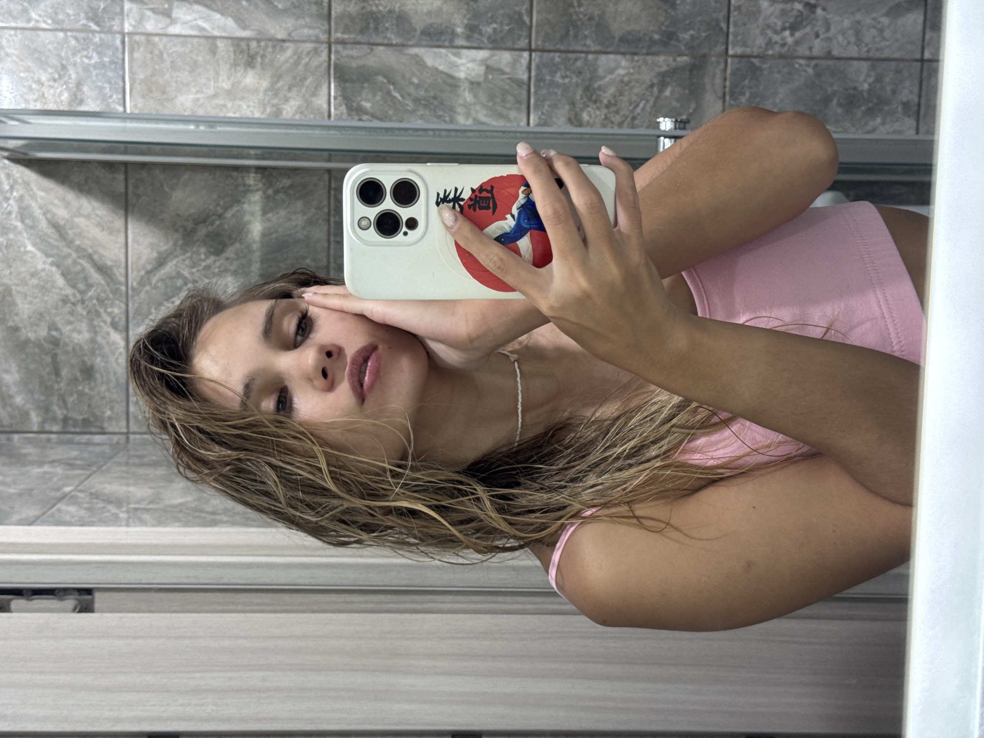 This rotated image captures a young woman taking a mirror selfie indoors. She is positioned horizontally due to the image's rotation, with her upper body visible. Her light brown, wavy hair appears damp, framing her face. She wears a pink tank top or crop top and a delicate white beaded necklace.
The woman holds a white smartphone with both hands, angled to capture her reflection. The phone case is distinctive, featuring a graphic of a red circle enclosing a stylized figure in blue and white, possibly depicting a martial artist or a character. To the left of this design, the Japanese or Chinese characters "蛛貓" (Zhū Māo), meaning "Spider Cat" or "Arachnid Cat," are visible. The phone's camera system, comprising three dark lenses, is clearly seen.
The background, reflected in the mirror, consists of grey, speckled tiled walls, suggesting a bathroom or a similar indoor environment. A light-colored shelf or ledge runs horizontally across the tiled surface. A white vertical strip on the far right indicates the edge of the mirror or a door frame. The lighting is uniform, characteristic of an indoor setting. The woman maintains a neutral expression, with her lips slightly parted. The location context provided is Chaikovsky, Russia.