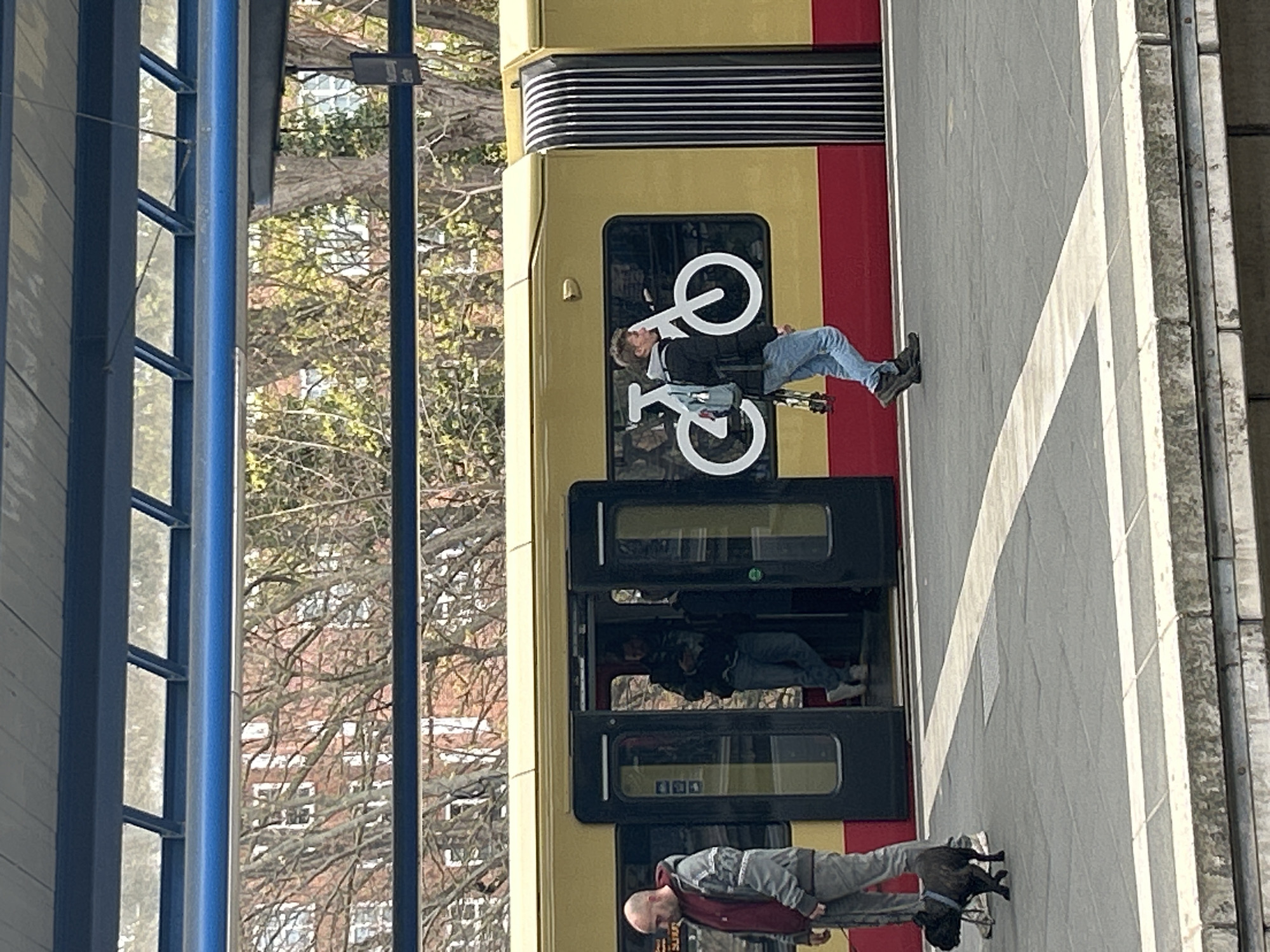 The image depicts a train station in Berlin, Germany. An S-Bahn train, identifiable by its yellow and red livery and bicycle symbol, is stopped at the platform. Two passengers are visible, one standing near the open doors, and another exiting the train. The train is at a stop adjacent to the platform. Architectural elements, including support beams, are evident on the left side. Trees with bare branches fill the backdrop.