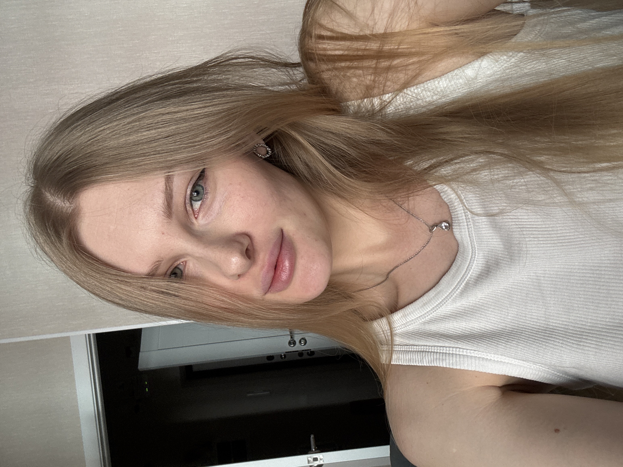 A young woman with fair skin, light blonde hair, and blue eyes is photographed indoors. She is wearing a white ribbed tank top and a delicate silver necklace. An earring is visible in her left ear. Behind her is a partially visible wall and what appears to be a dark-colored appliance or electronic device, possibly a television or monitor, mounted on a light-colored surface. There are no indications to determine the specific location in the city of Perm, Russia.