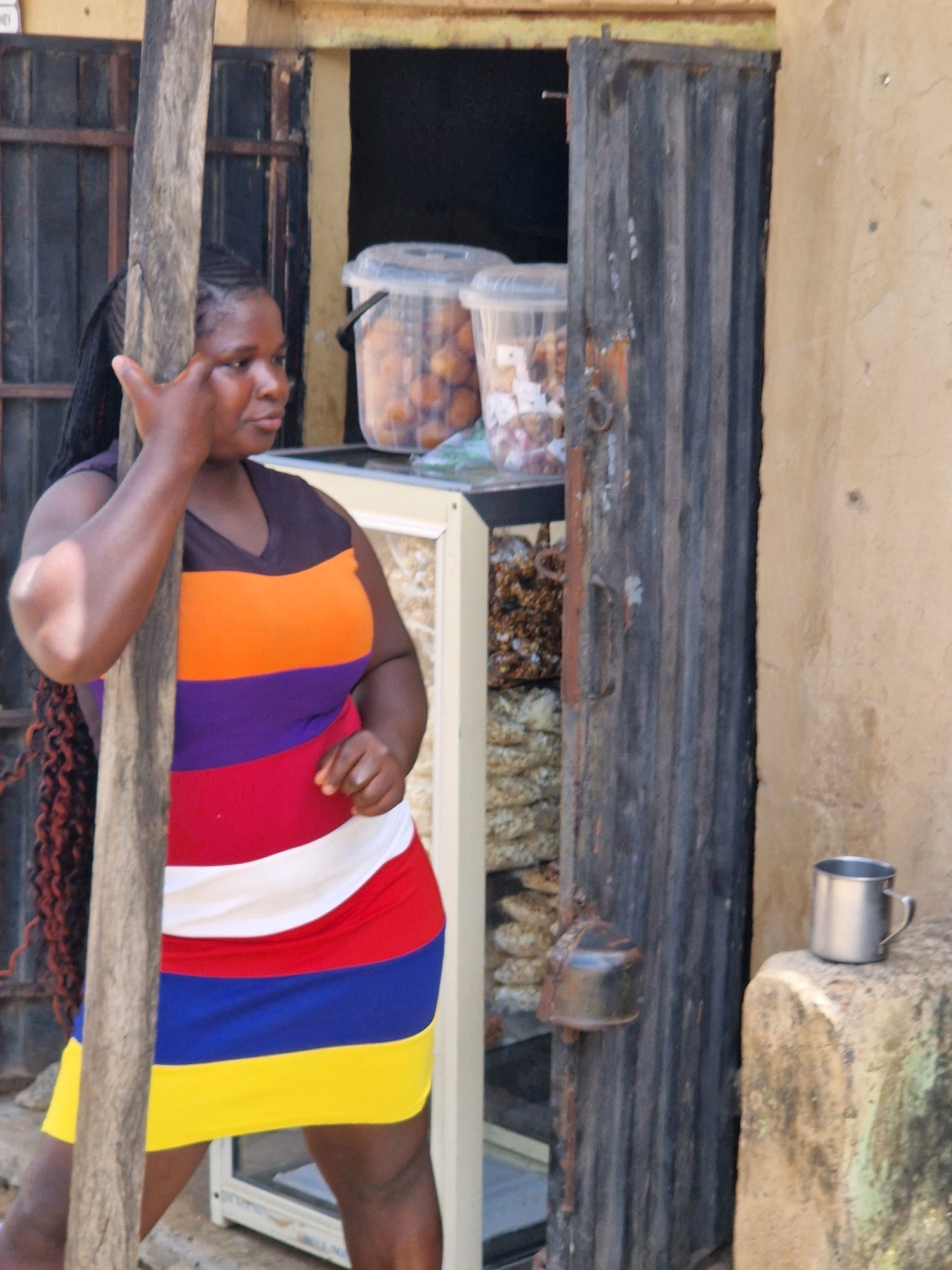 A woman with dark braided hair and a multi-colored striped dress stands in front of a weathered shop. The dress features horizontal stripes of purple, orange, red, white, blue, and yellow. Her left arm is raised, with her index finger pointing towards her forehead. Behind her, a cooler is visible, containing translucent containers filled with small, round, brown items, possibly food. A metal door, possibly corrugated iron, is ajar to her right. Next to the door, a silver mug rests on a rough, beige stone. The background appears to be an outdoor, urban setting with a textured wall.