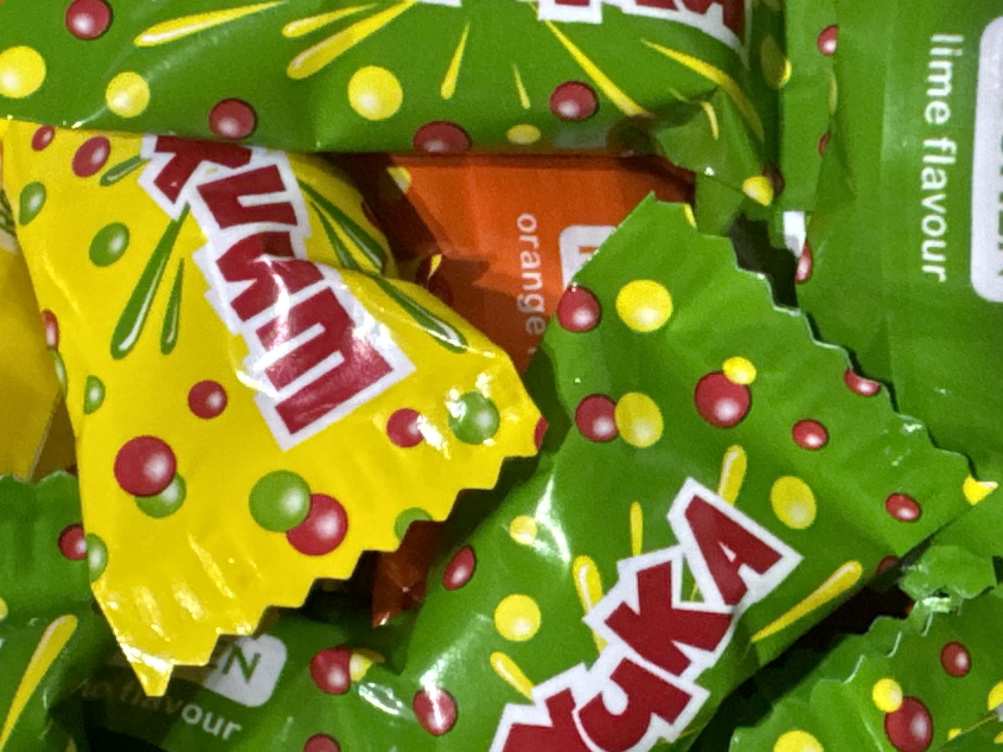The image is a close-up, high-angle, macro shot of several brightly colored candy wrappers, predominantly green and yellow with red and white lettering. The wrappers are crumpled and overlap each other, suggesting they are likely discarded or in a pile. Some of the wrappers have circular patterns of red and yellow dots, resembling candies themselves. Text is visible on some of the wrappers, including what appears to be "orange" in white lettering on an orange wrapper, and "lime flavour" in white lettering on a green wrapper. The overall impression is one of a messy collection of confectionery packaging. There are no people, animals, or environmental elements of Waterford, Ireland visible in this image.