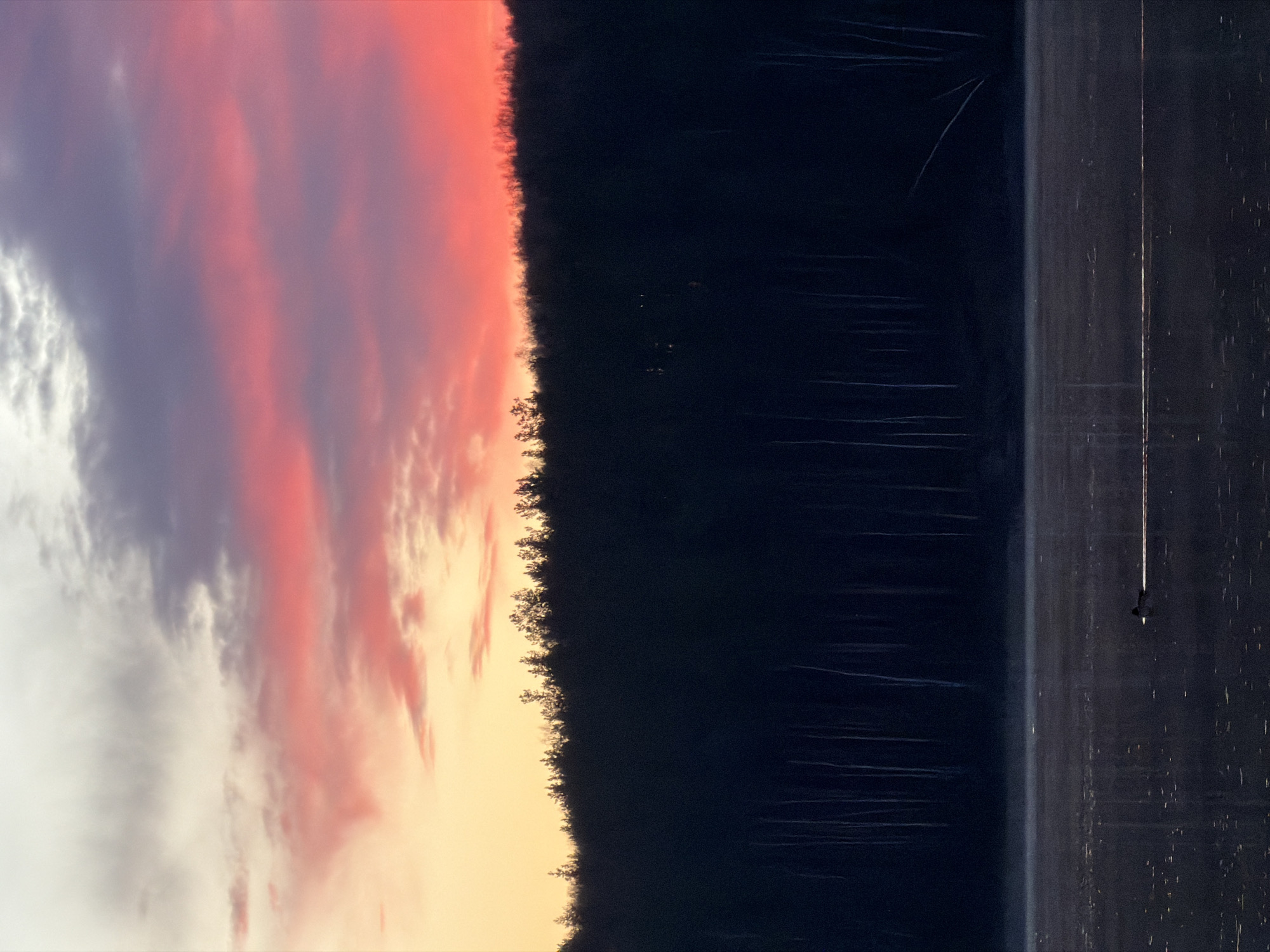 A vertical shot depicts a body of water reflecting trees and a colorful sky. At the bottom right, a person is walking on the water's edge. The sky is filled with white, grey, purple, pink, orange, and yellow hues. A dark forest line separates the sky from the water. The shot appears to be taken during sunrise or sunset. Based on the location provided, it is likely taken on the Kama River near Chaykovsky, Russia.