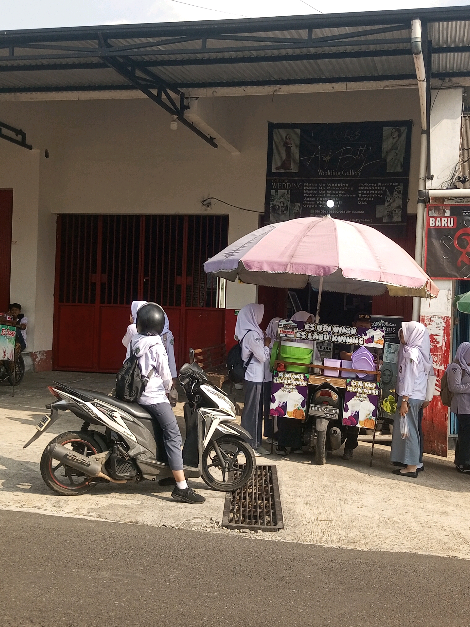 In Krasak, Indonesia, a street scene unfolds near a building with a corrugated metal awning and red double doors. A food cart, adorned with colorful signage featuring pumpkins and purple fruit, is parked on a concrete sidewalk. The cart is topped with a large pink and white umbrella providing shade. Several young people, likely students wearing white uniforms and hijabs, are gathered around the cart. One individual, wearing a black helmet, is seated on a white and black motorcycle in the foreground, facing the cart. Another student stands near the cart holding a plastic bag, while others are visible in the background. The building behind the cart has a banner that reads "Ayu Betty Wedding Gallery," listing various services. A red sign with "BARU" (new) is visible to the right. The overall environment appears to be a commercial area with moderate pedestrian activity.