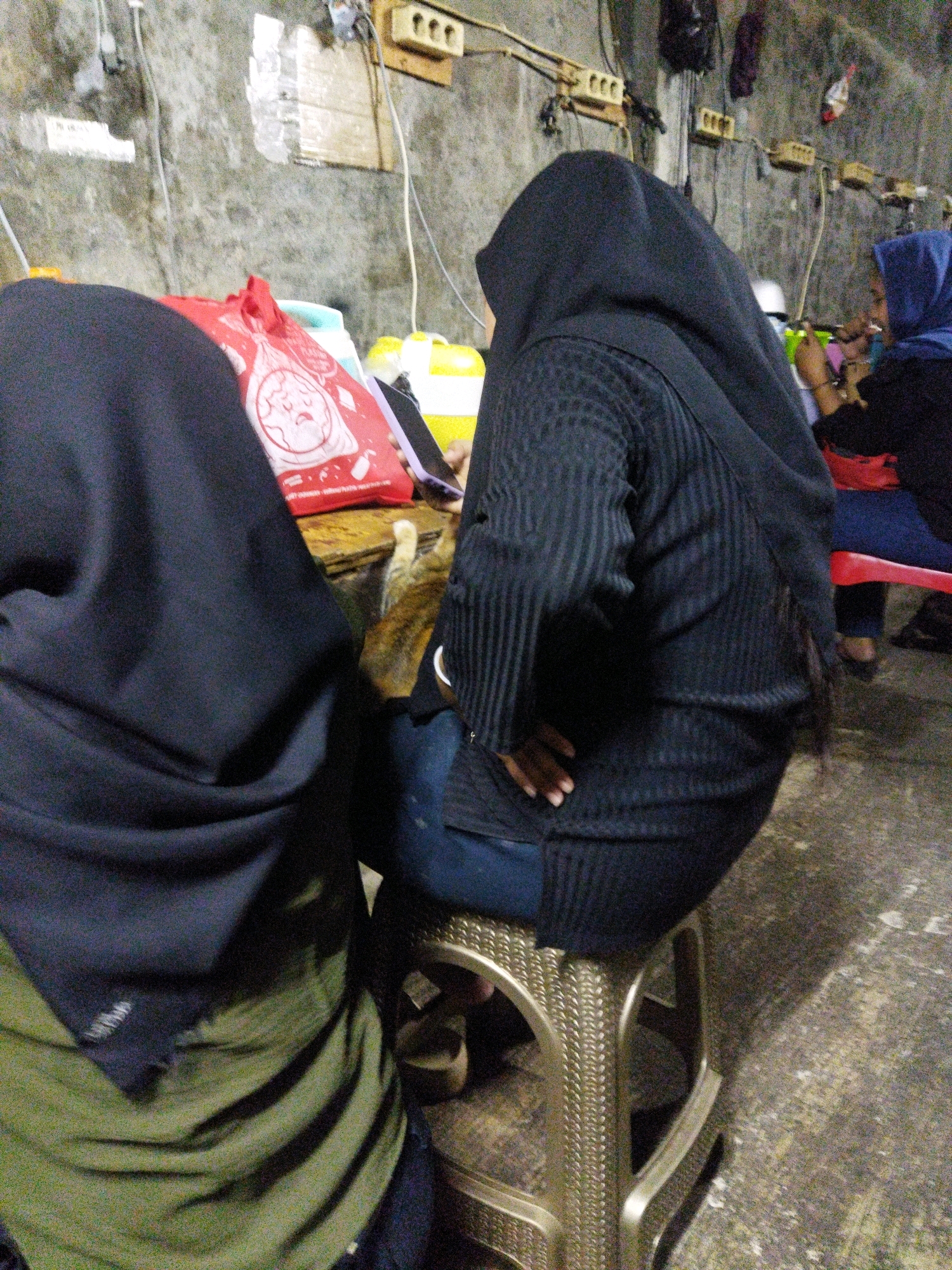 The image depicts an indoor scene, likely a casual eatery or roadside stall, in West Jakarta, Indonesia. Three individuals, two women wearing hijabs and one partially visible, are seated. The woman in the foreground, facing away from the camera, is dressed in a black hijab and a striped black sweater over blue pants. She is seated on a woven plastic stool. To her left, another individual, also wearing a dark hijab, is visible from the back, with a green and black garment. To the right, a woman wearing a blue hijab is seated, and appears to be smoking. A red bag with a circular graphic is placed on a table in front of the woman in the foreground. Behind the individuals, a concrete wall with electrical outlets and wiring is visible. The ground is concrete. The overall atmosphere suggests a simple, communal setting.